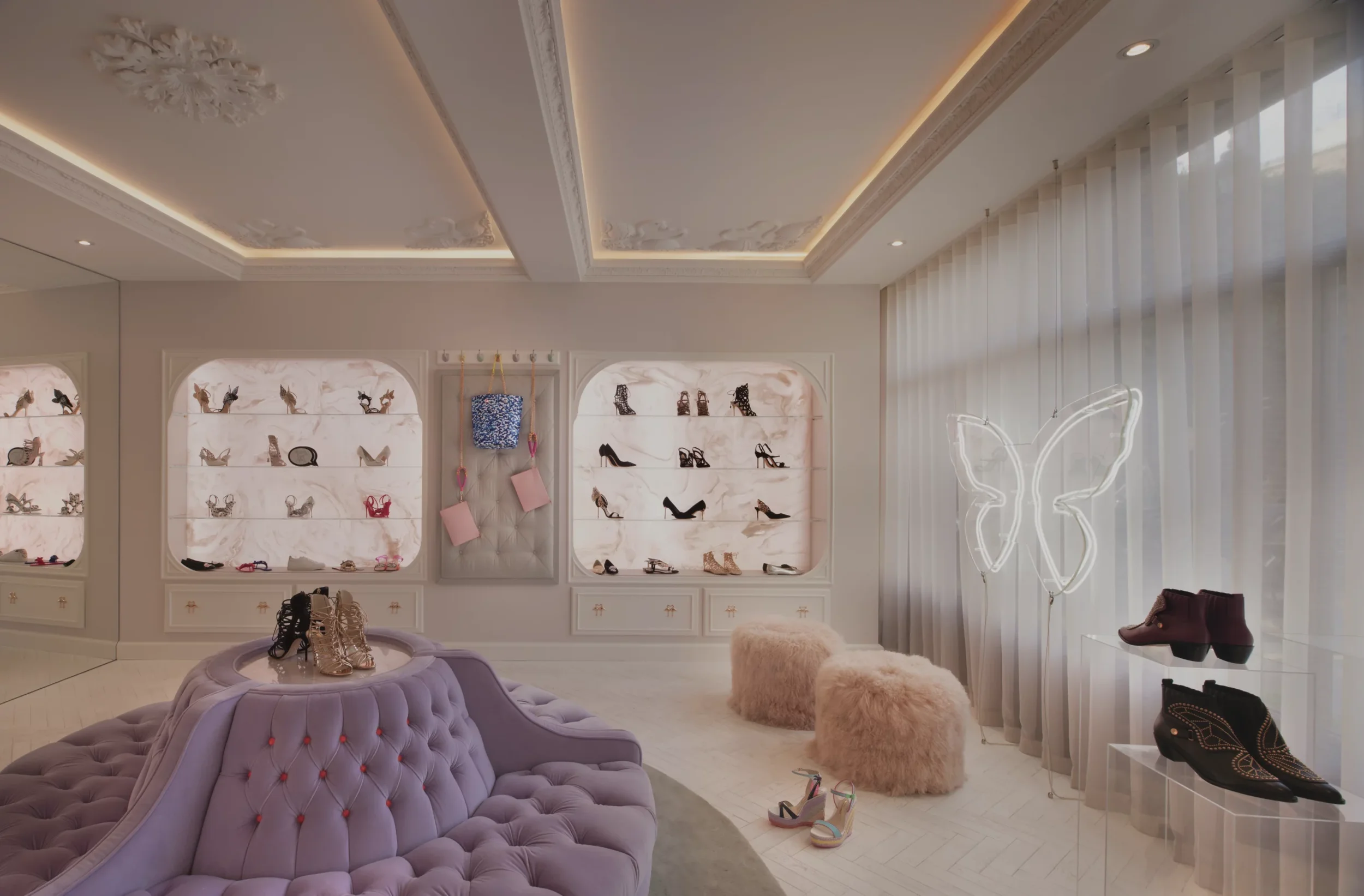 Interior of a shoe boutique with display shelves of high-heeled shoes, a neon butterfly sign, plush ottomans, and decorative lighting.