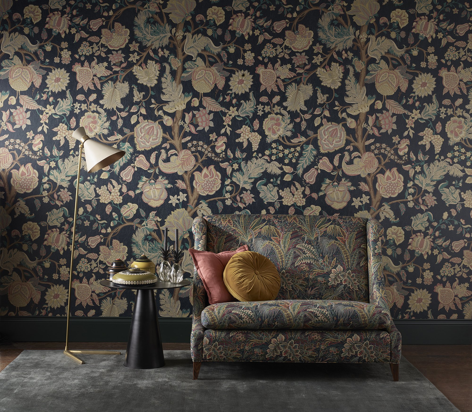 A living room corner with Liberty London floral patterned wallpaper, a modern floor lamp, a side table with decorative vases, and a floral upholstered sofa with a pink pillow and a gold circular pillow.