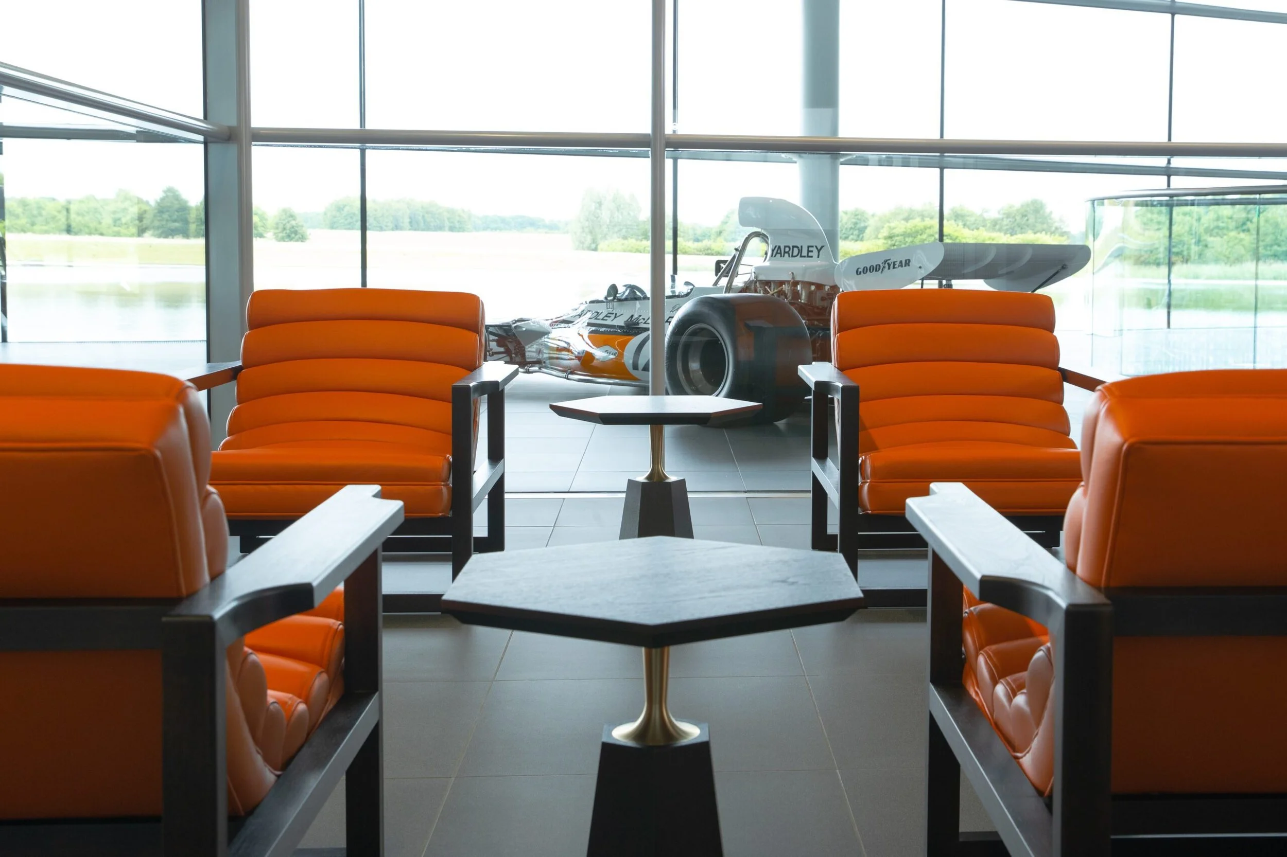 Inside view of an airport lounge holding area with orange chairs and a small table, looking out large glass windows at a scenic lake and green landscape. A racing car is displayed behind the glass.
