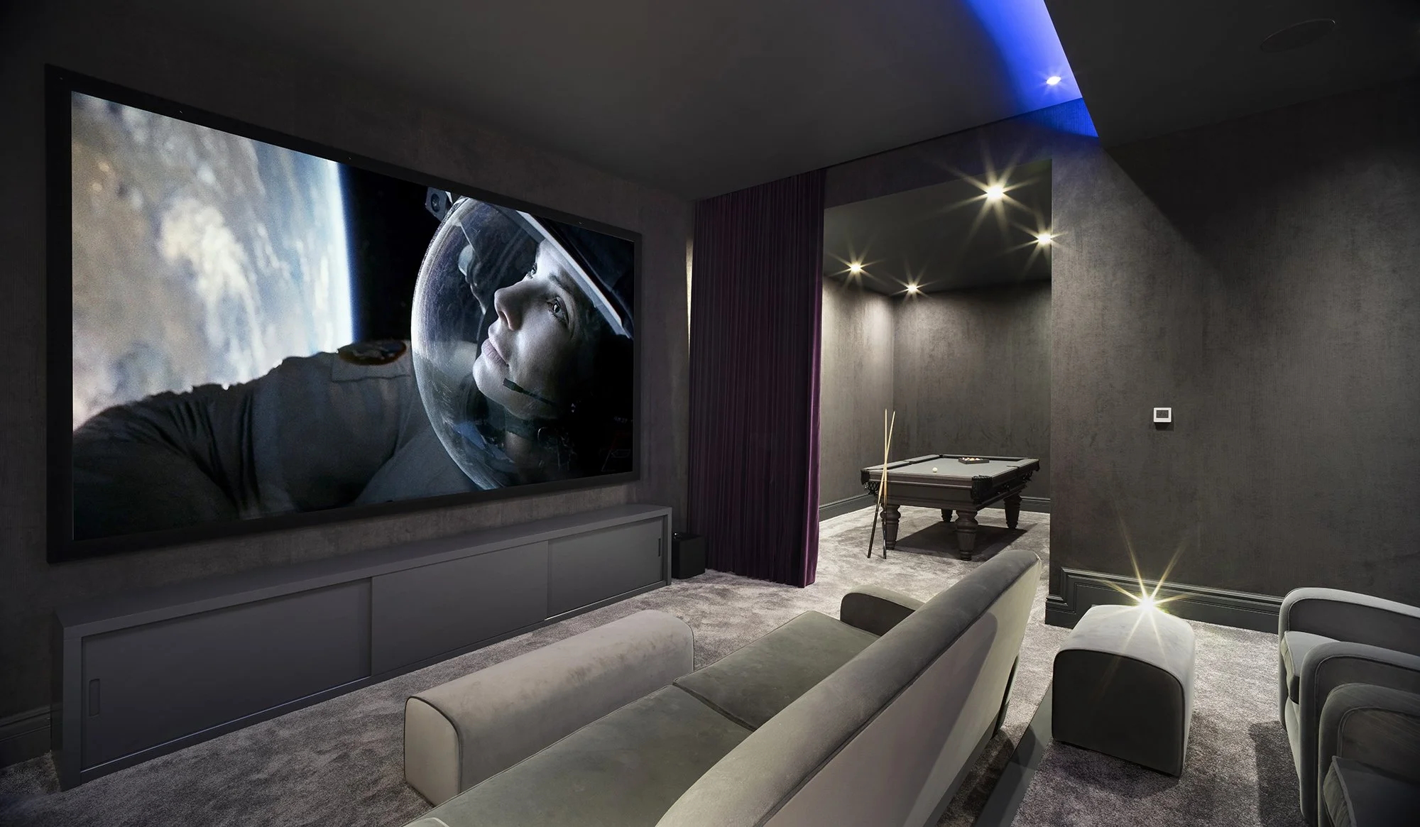 A modern home entertainment room with a large flat-screen TV showing an astronaut in space. The room has gray walls, a gray carpet, a gray sofa, and a pool table in the corner. There are blue and warm ceiling lights, and a purple curtain partially covering the doorway.