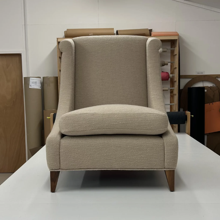 A beige upholstered armchair with wooden legs, sitting on a white surface in a workshop or storage area with rolls of fabric and other materials in the background.
