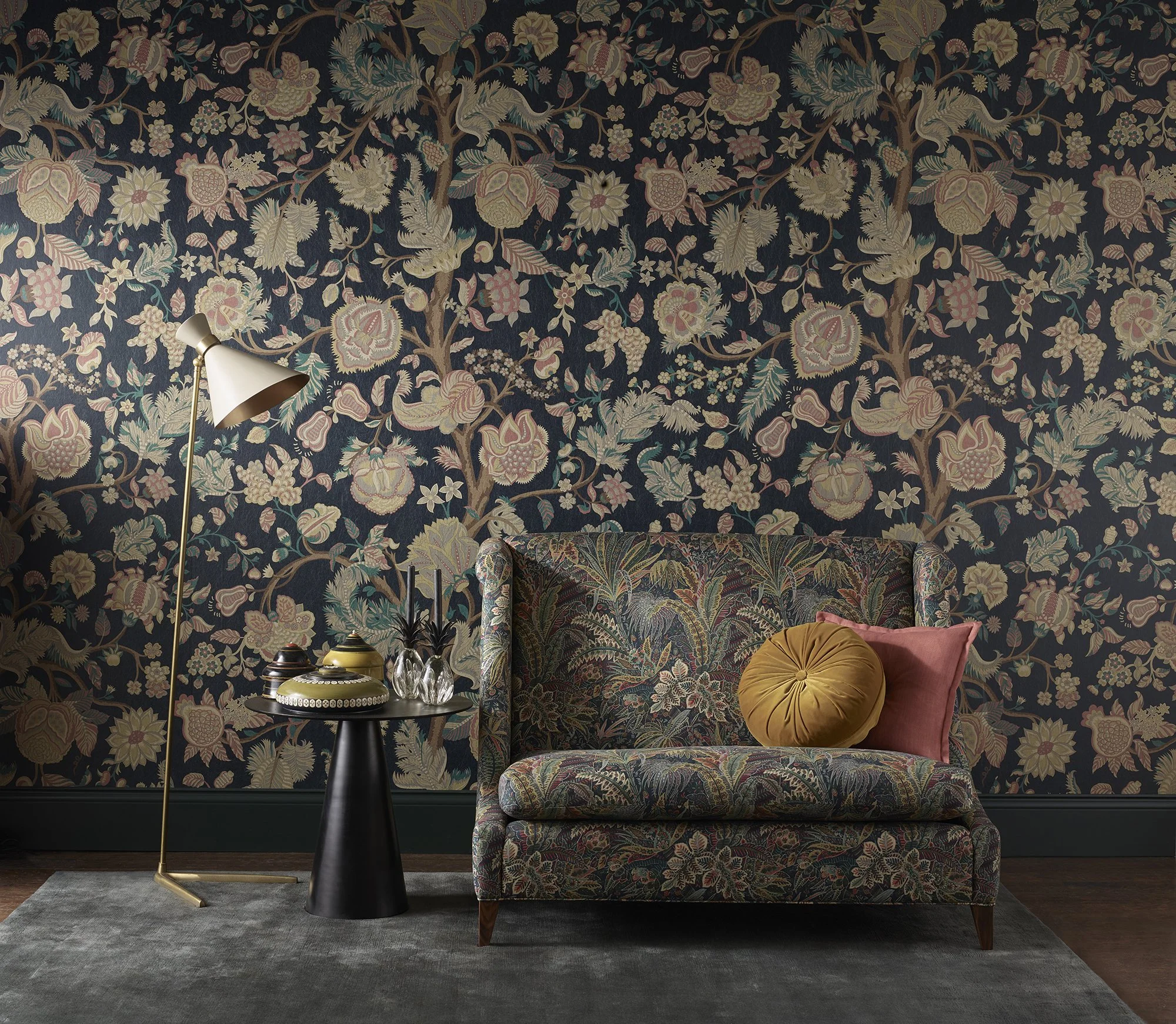 A living room with a dark, floral-patterned wallpaper, a floral upholstered sofa, a mustard-colored round pillow, a black side table with decorative bowls, a white floor lamp, and a gray rug.
