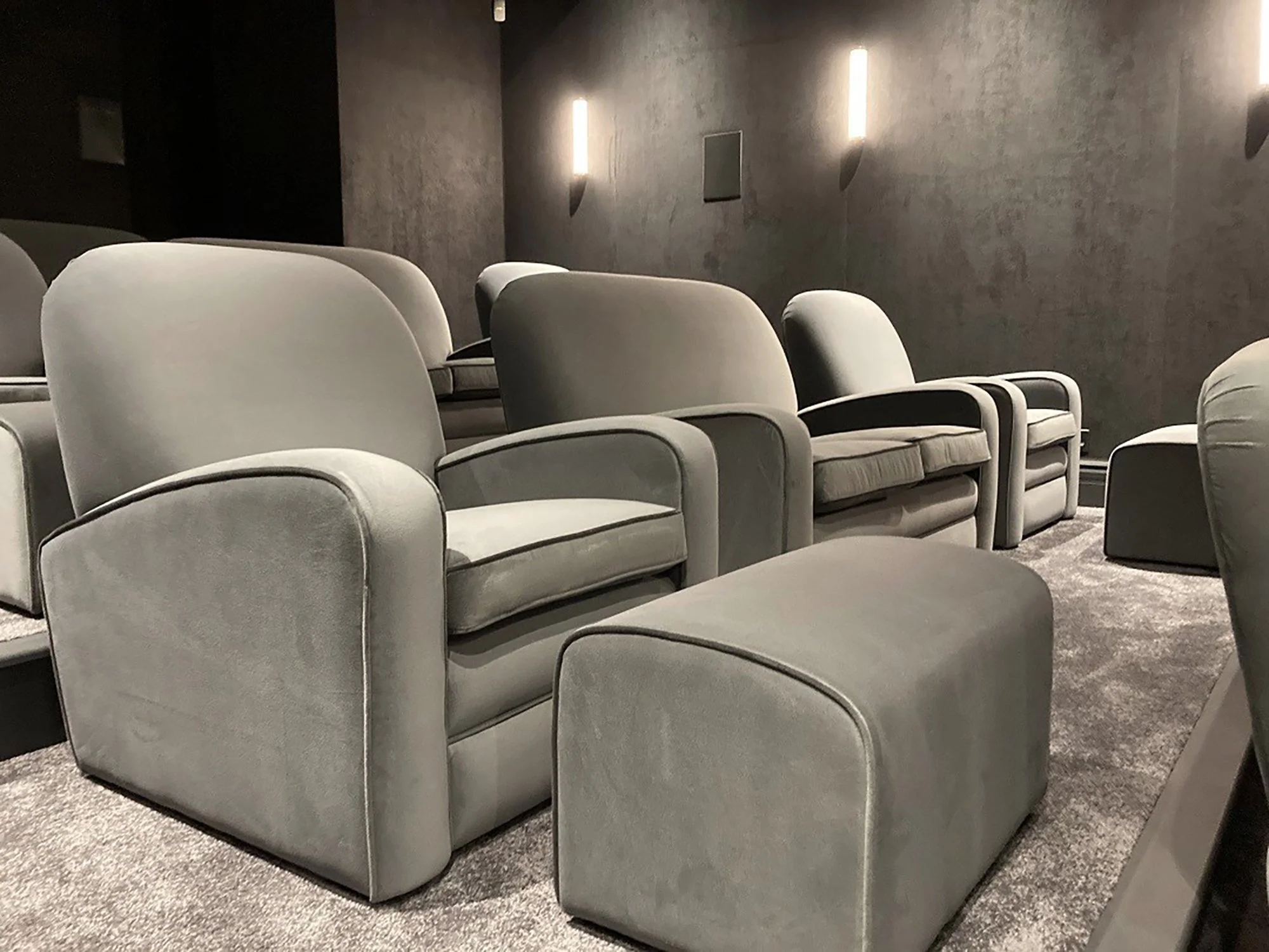 A row of gray cushioned lounge chairs in a dark, minimalist room with textured gray walls and vertical wall-mounted lights.