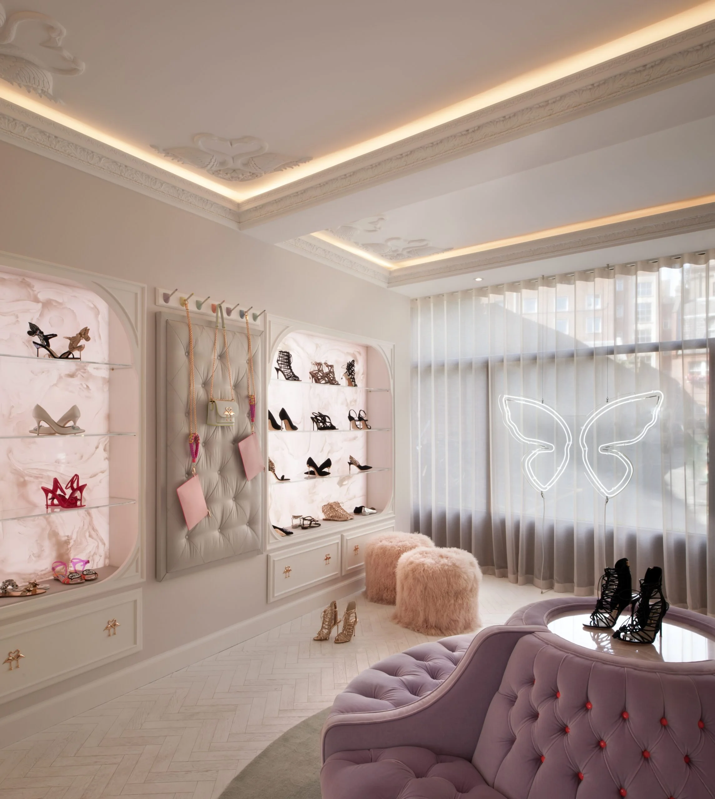 A boutique store interior with display shelves of high-heeled shoes and handbags, a tufted pink and purple sofa, soft plush ottomans, and large windows with sheer curtains, illuminated by natural light and ceiling lighting.