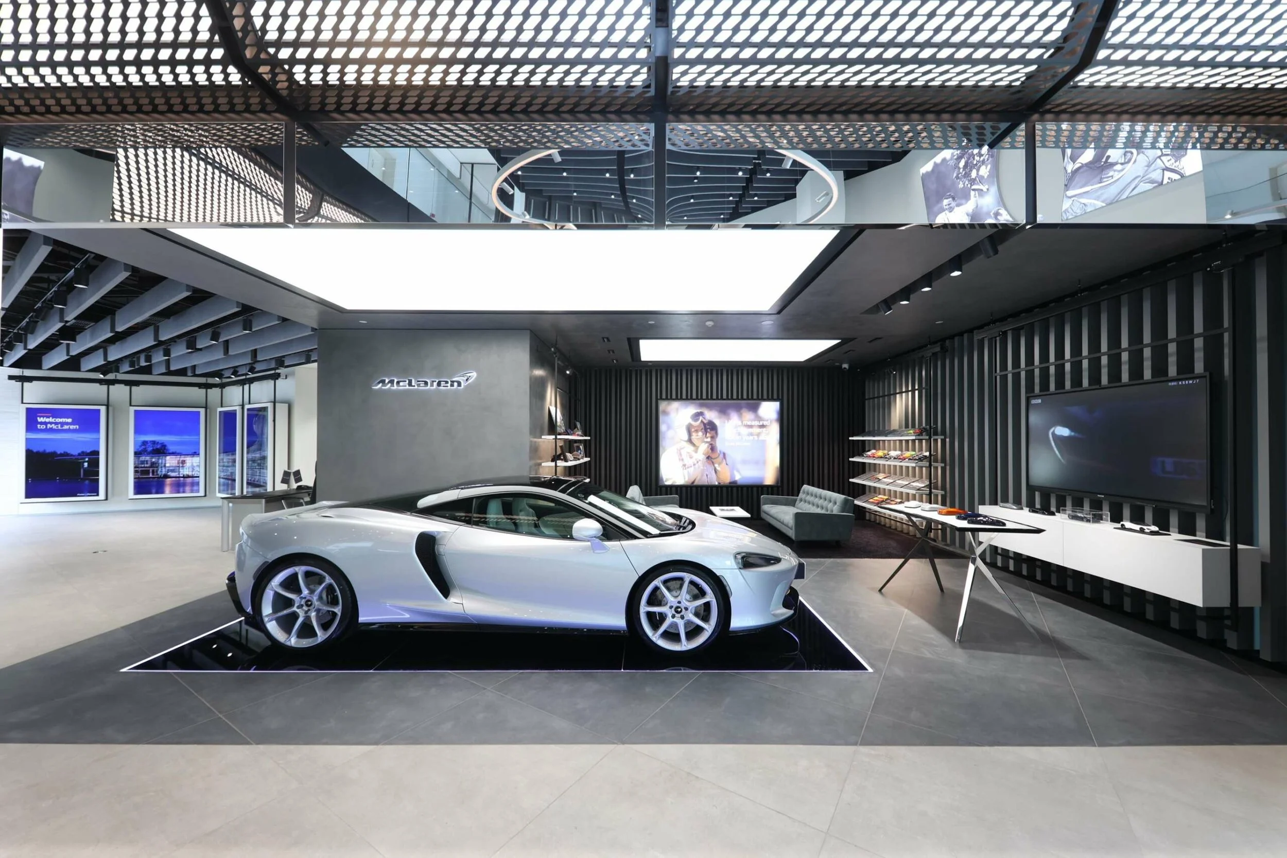 A white sports car displayed inside a modern McLaren showroom with dark and metallic interior design, large digital screens, and a seating area.