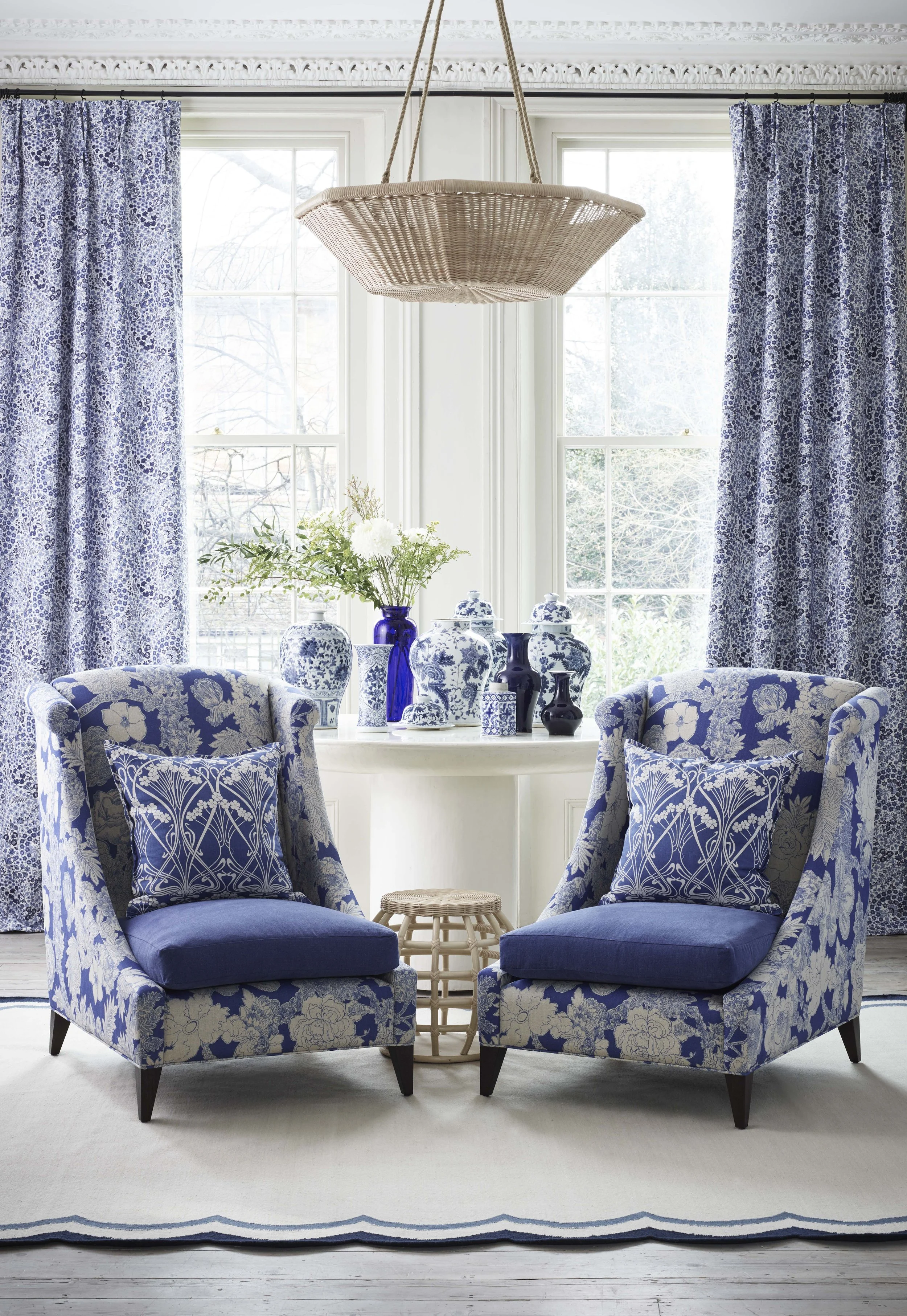 Interior of a bright living room with two blue floral Liberty Fabric patterned Vegas Lounger  armchairs, a round table between them adorned with blue and white vases.