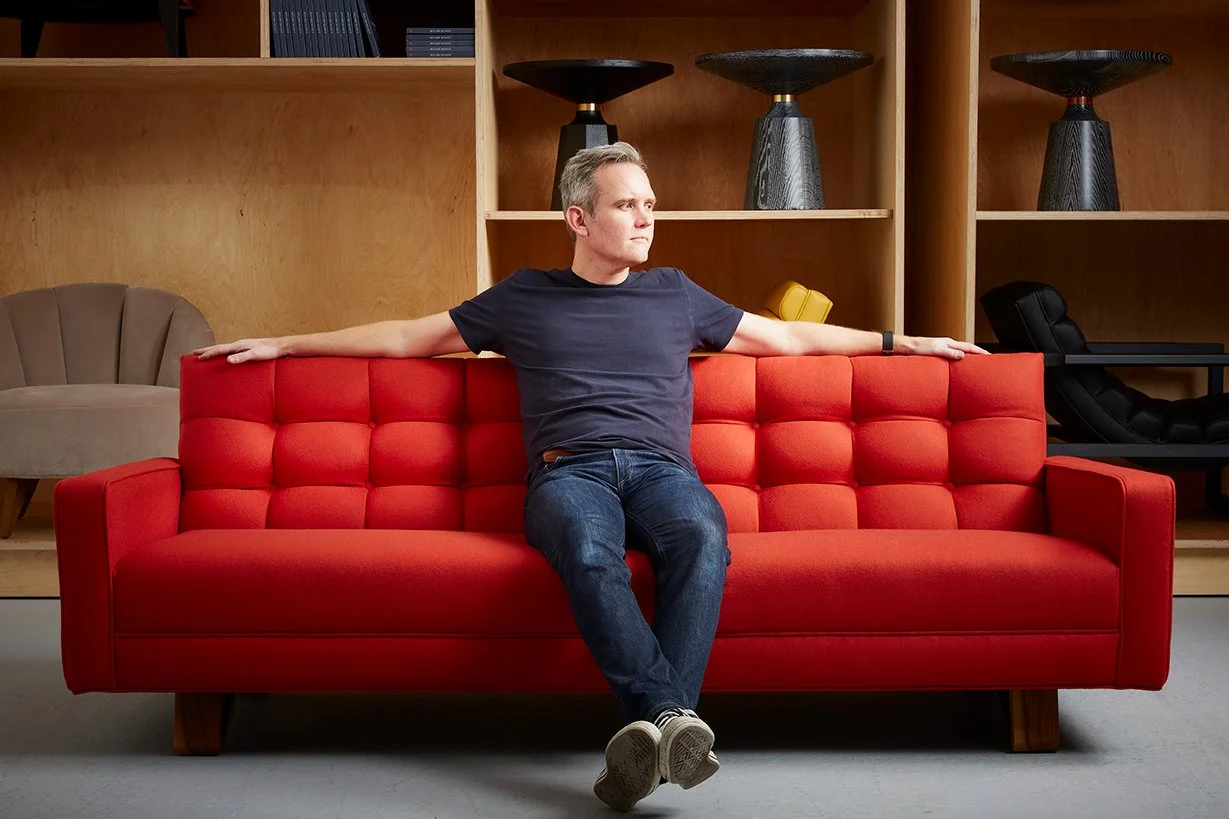 A man in a dark t-shirt and jeans sitting on a bright red sofa with arms outstretched, in a room with wooden shelves and decorative objects.
