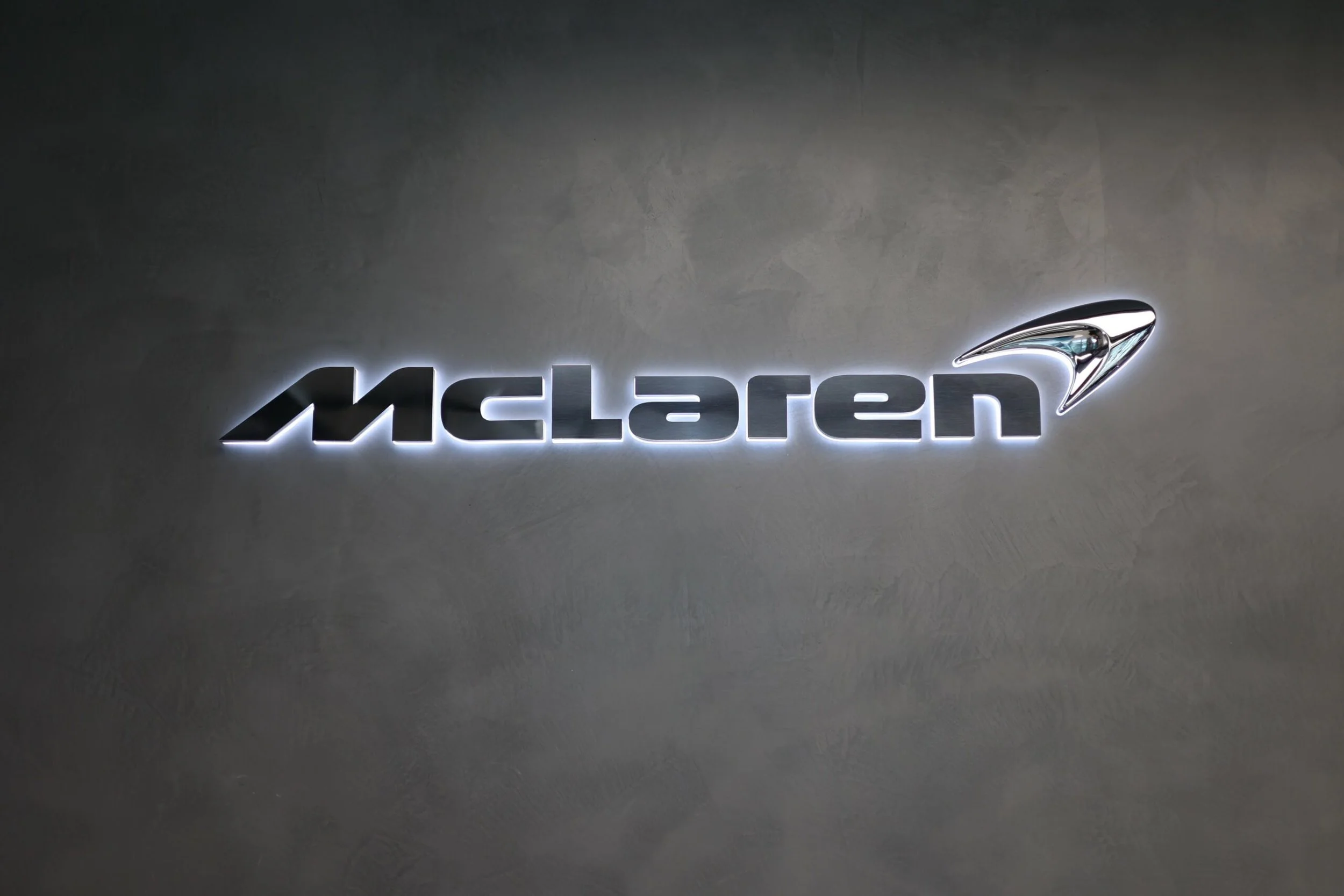 Backlit McLaren logo on a gray wall with a sleek, metallic emblem of a speedster above the text.