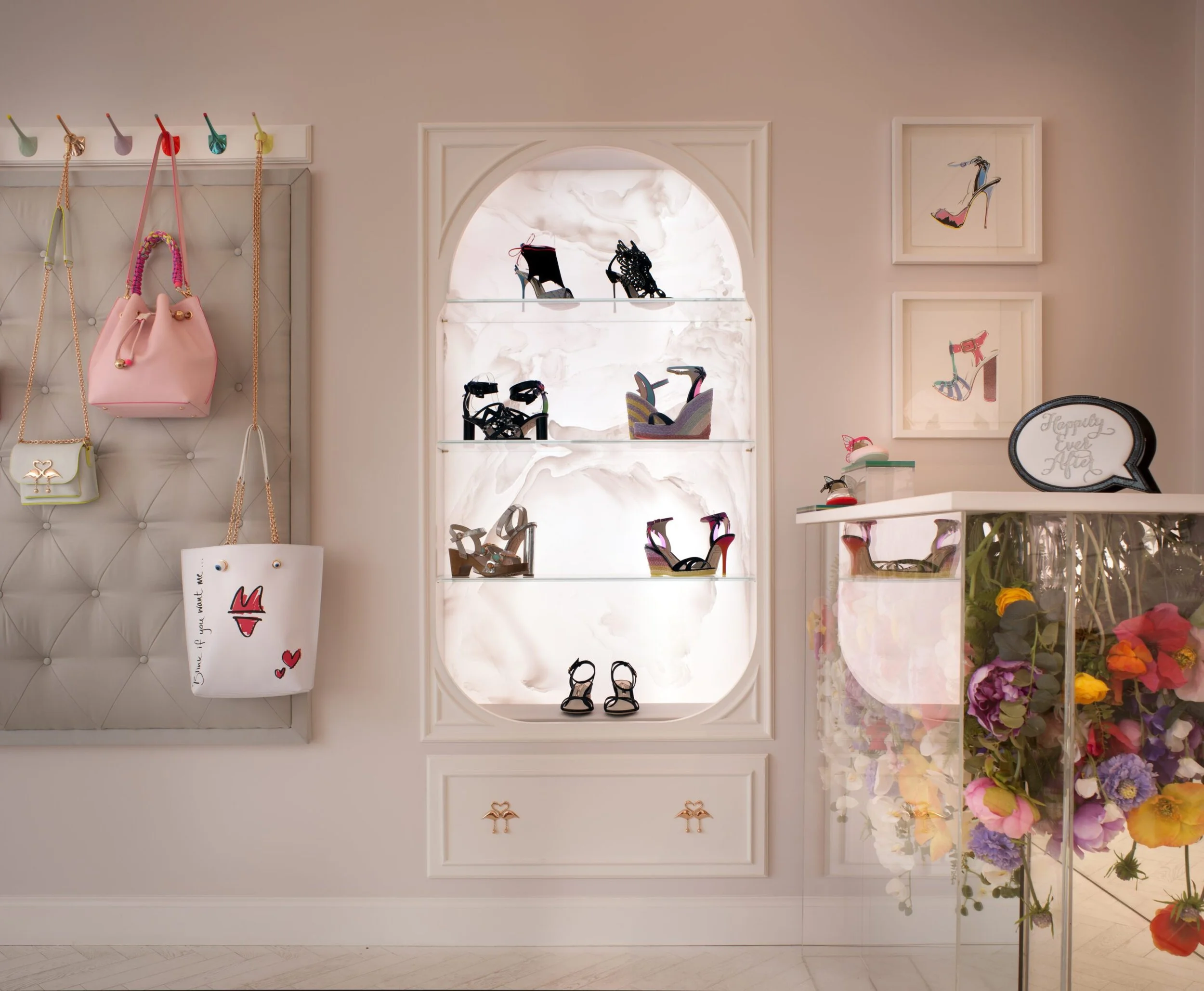 Display of high-heeled shoes on shelves, framed shoe illustrations on the wall, and handbags hanging on a tufted wall panel, with floral arrangements on the right side.
