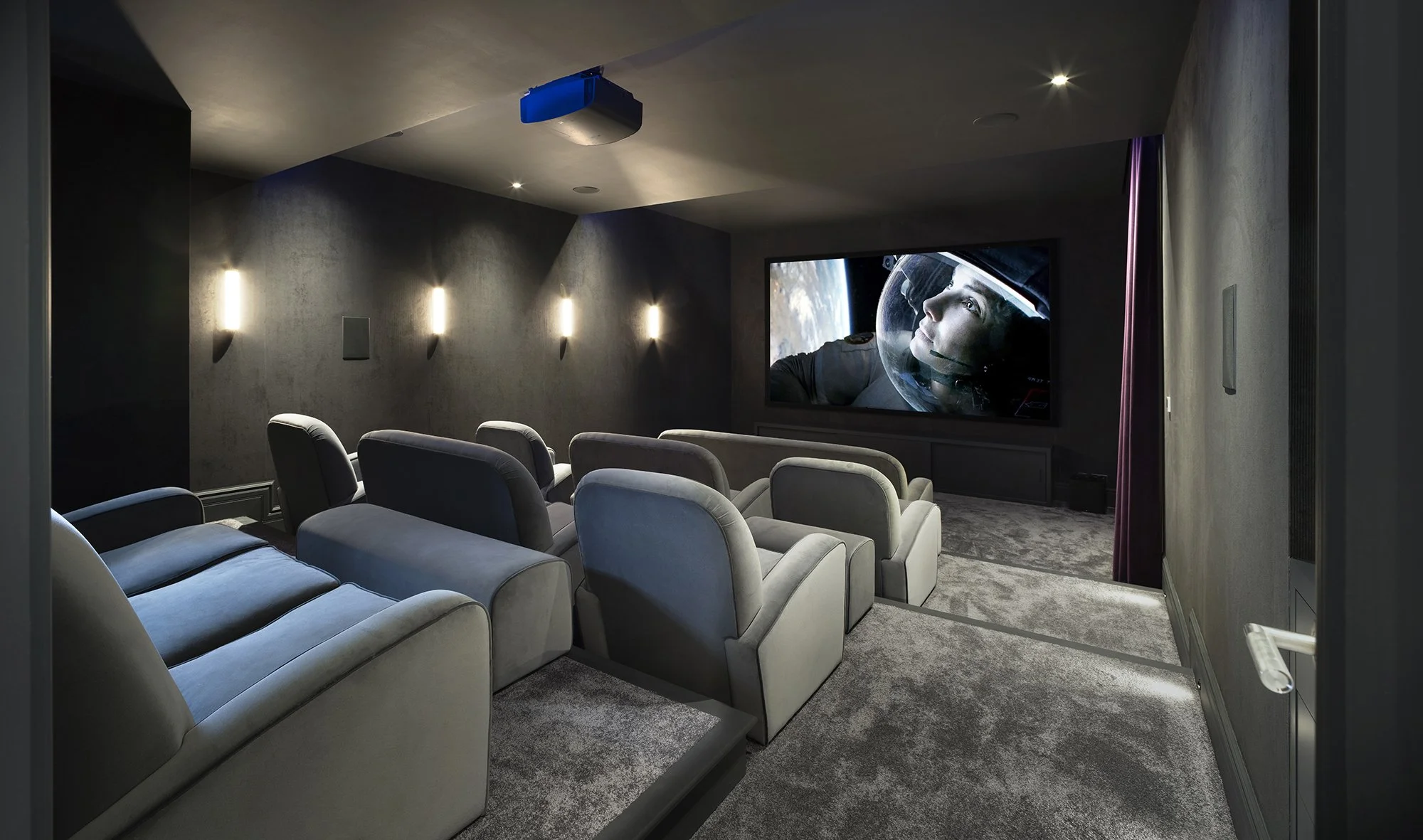 Home theater with multiple beige recliner chairs, a large screen displaying an astronaut, and ambient lighting on dark walls.