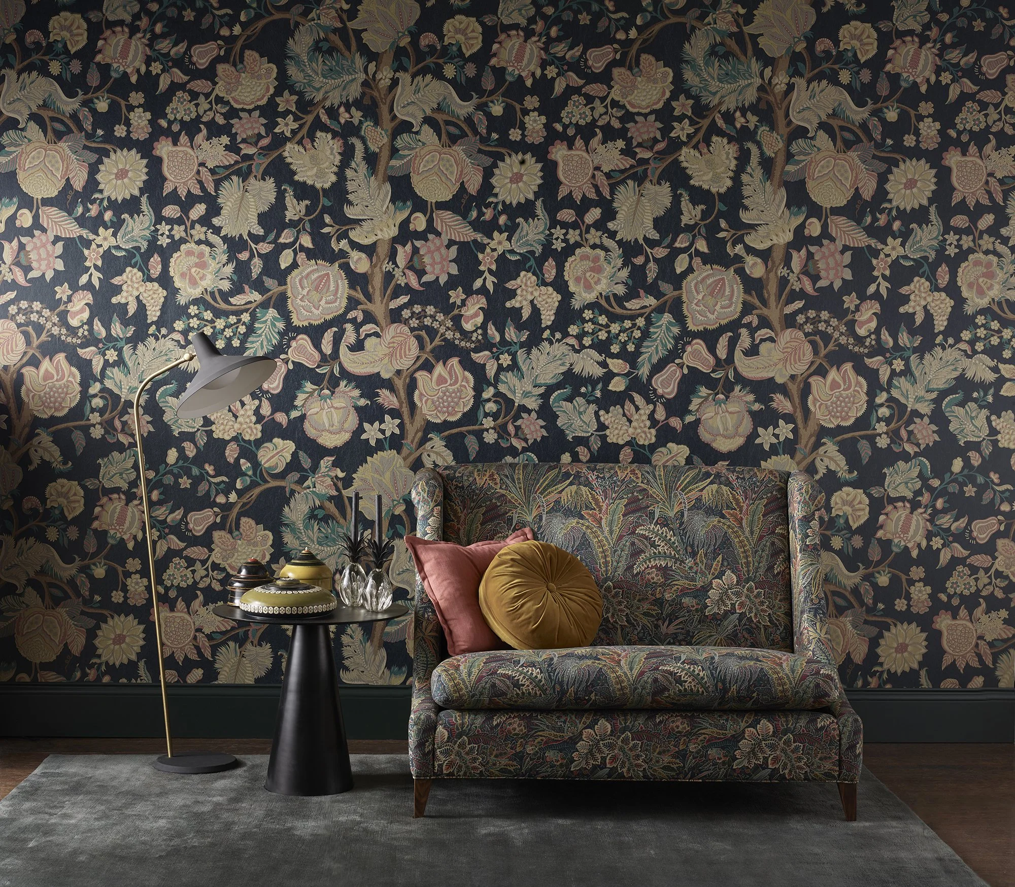 A living room with a dark floral wallpaper featuring birds, flowers, and leaves. A matching floral armchair is in front of the wallpaper. To the left, a black side table holds decorative vases, and a white floor lamp with a curved stand is positioned beside the table.