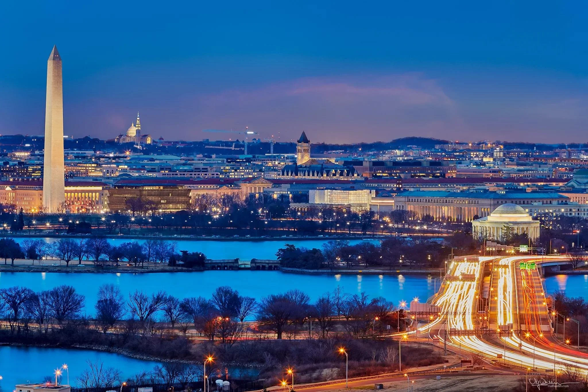 Nighttime cityscape of Washington D.C. featuring the Washington Monument, the Capitol Building, and the Smithsonian Institution with a river in the foreground and a busy highway with light trails.