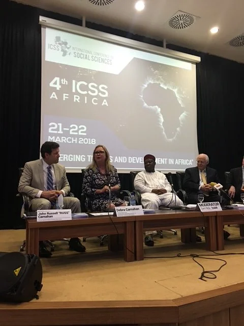 Panel of four people sitting at a conference table on stage, with a large screen behind them displaying text about the 4th ICSS Africa conference, held March 21-22, 2018, covering topics on emerging issues and development in Africa.
