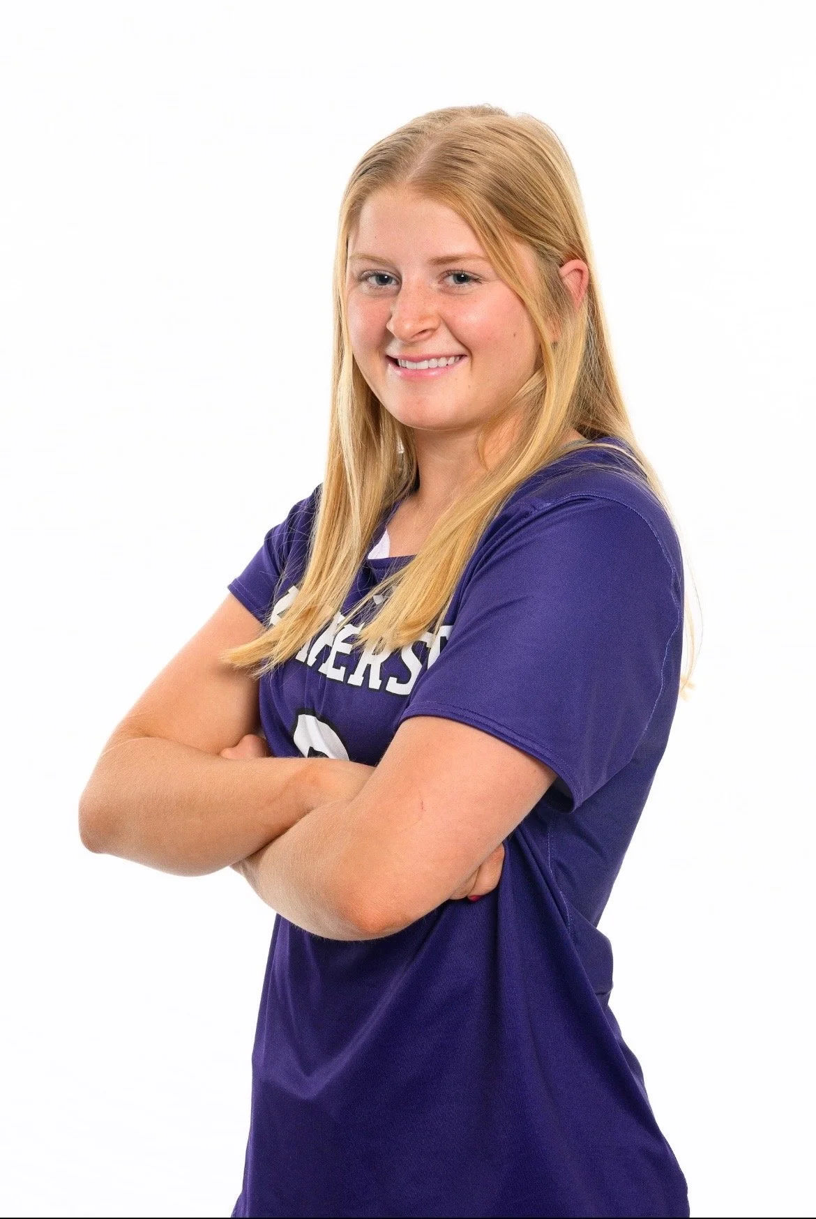 A young woman with long blonde hair, wearing a purple sports jersey with crossed arms, looking at the camera and smiling against a white background.
