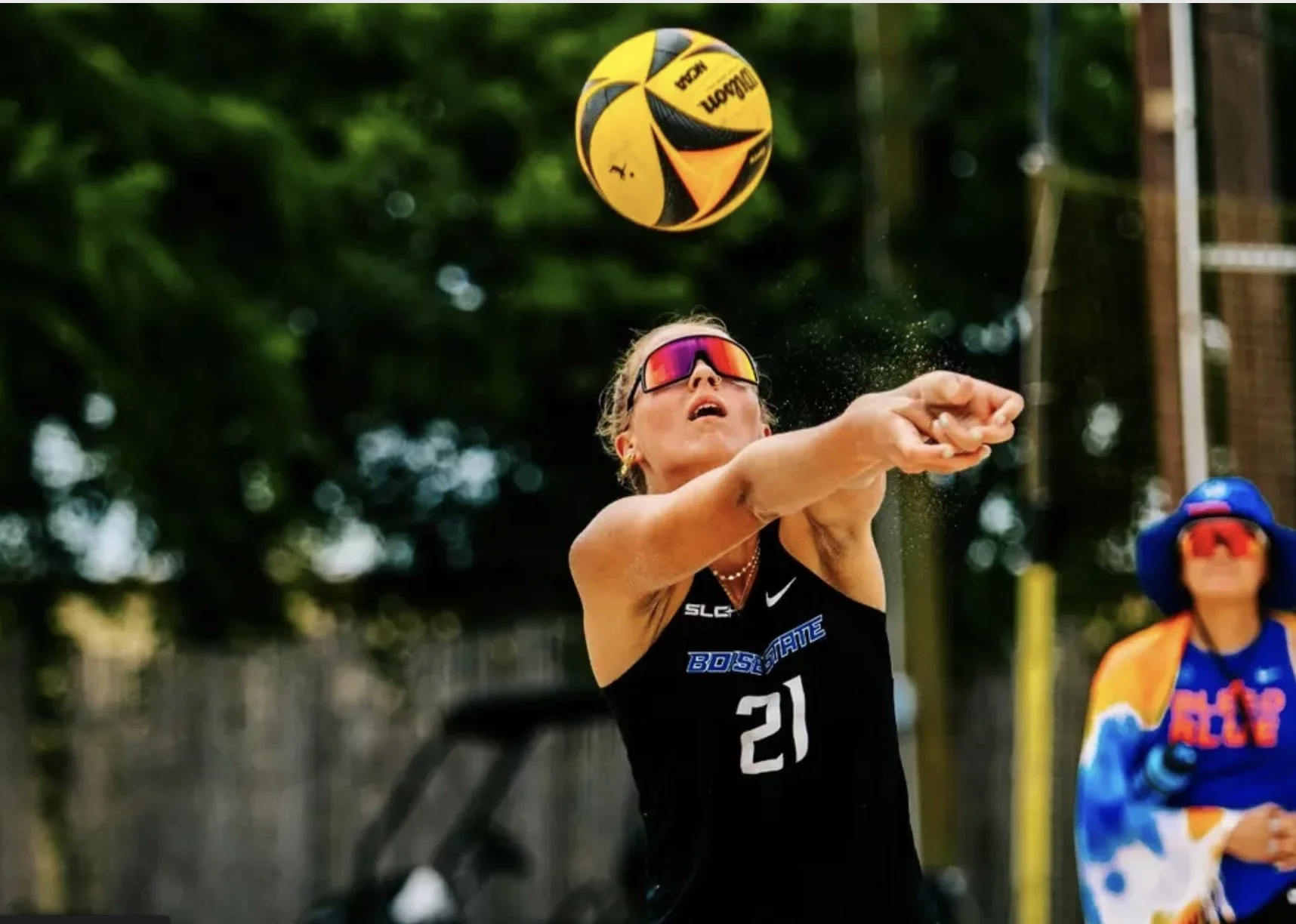 A female beach volleyball player wearing sunglasses, a black athletic top with the number 21, and a necklace, is preparing to hit a yellow and black volleyball during a match outdoors with a blurred background of trees and a fence, and a referee or another player in colorful attire in the background.