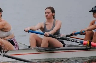 Three women rowing a boat on a body of water, two women are actively rowing with blue oars, one woman appears to be resting or waiting.