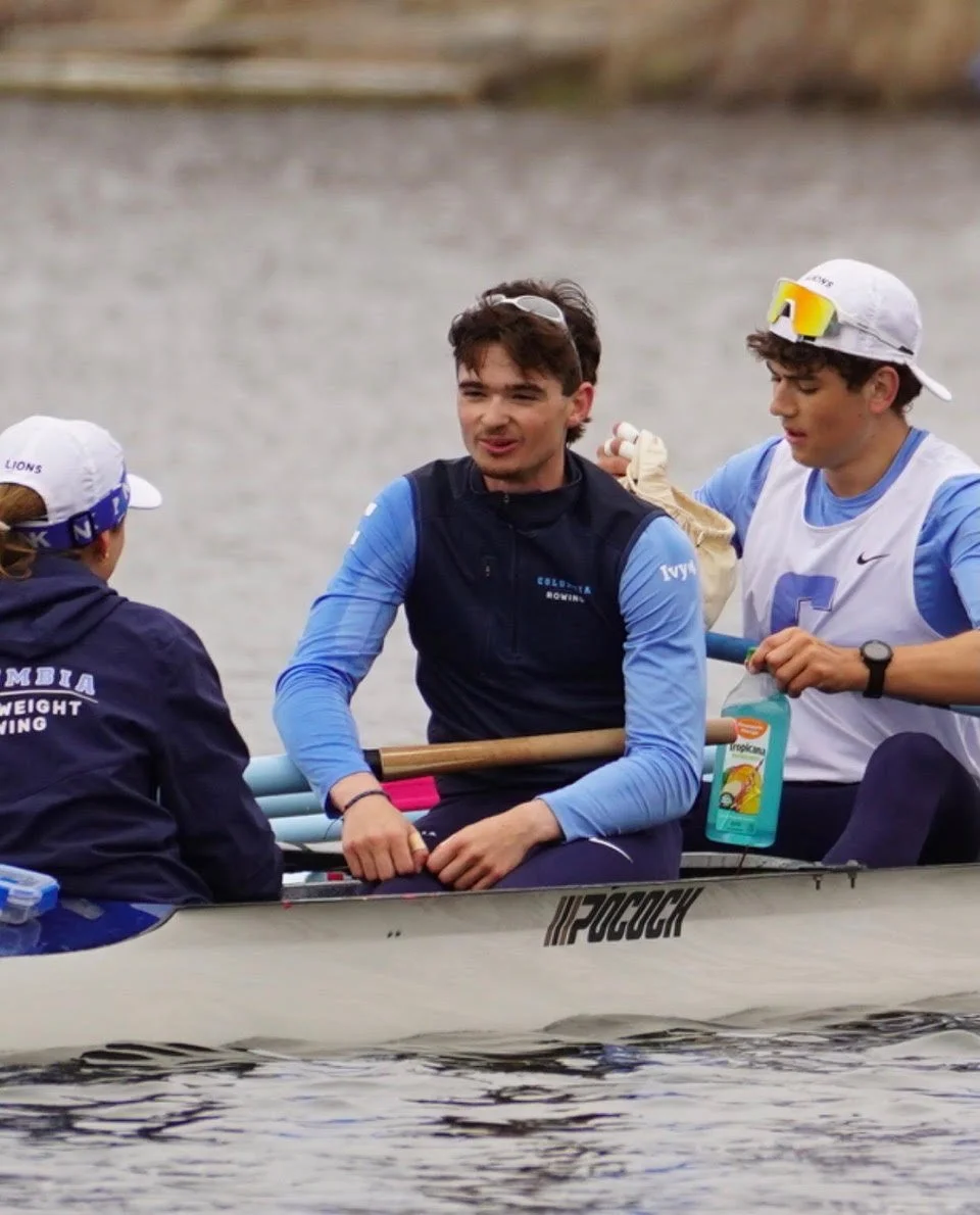 Three young men in a rowing boat by a body of water, one sitting and the other two standing, with one holding a paddle and the third adjusting equipment, all wearing athletic gear.