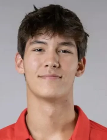 Close-up of a young man with short, dark hair, wearing a red shirt, smiling slightly.