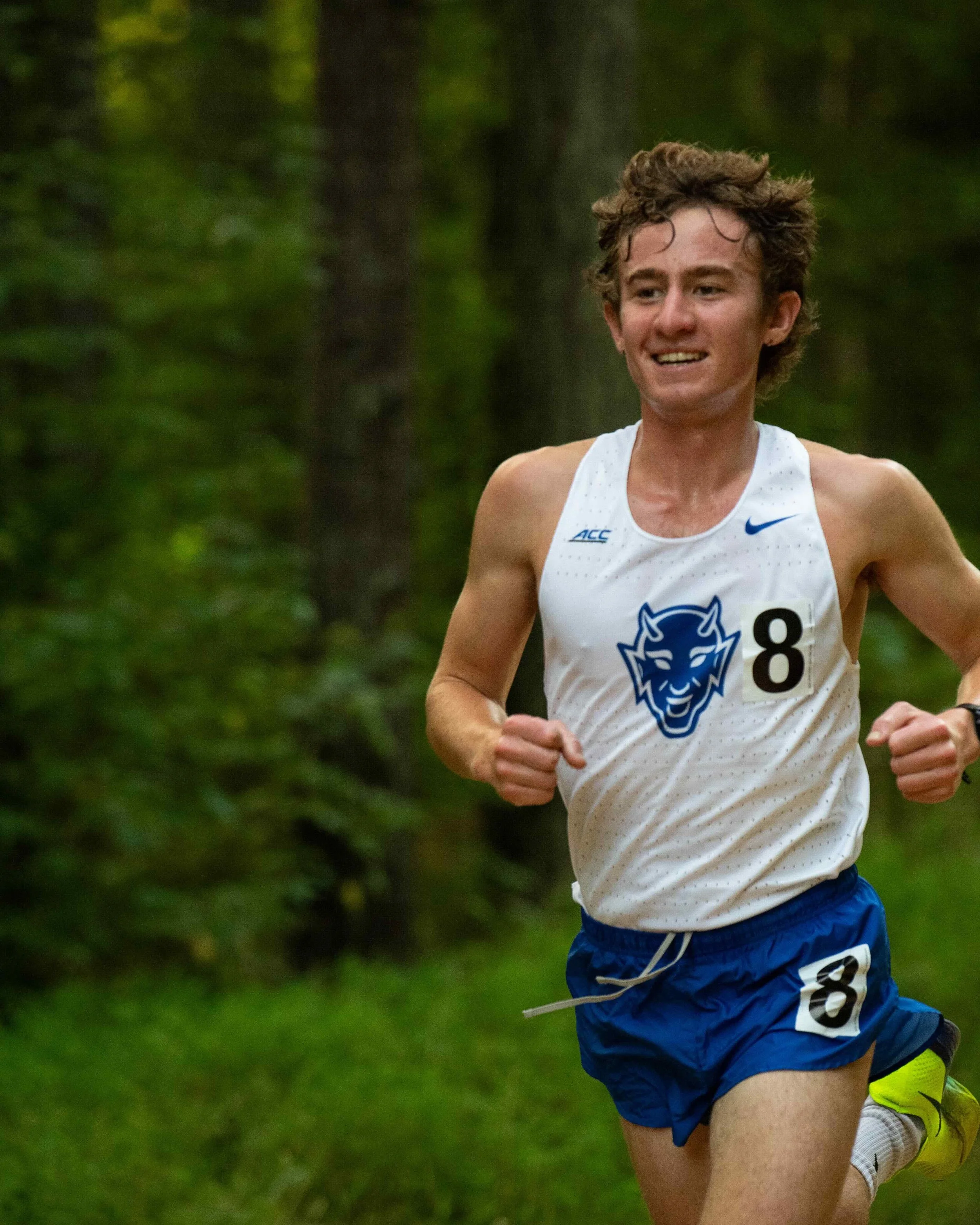 A male cross-country runner with curly brown hair, wearing a white tank top with blue logos, blue shorts with a number 8, and yellow shoes, running through a wooded area.