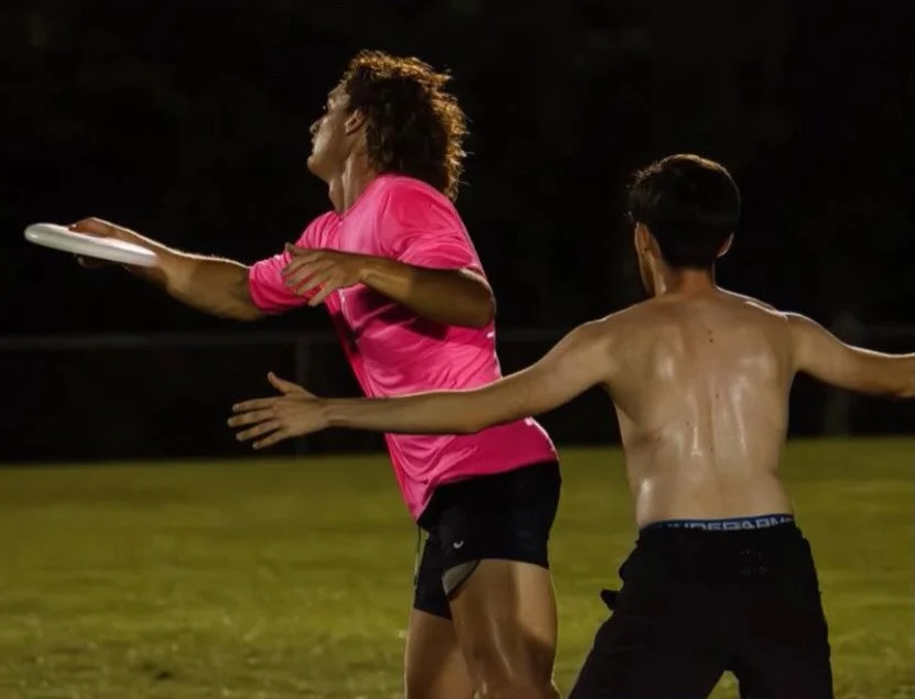 A woman in a pink shirt and black shorts is playing frisbee on a grassy field at night, with a shirtless boy reaching out to catch the frisbee.