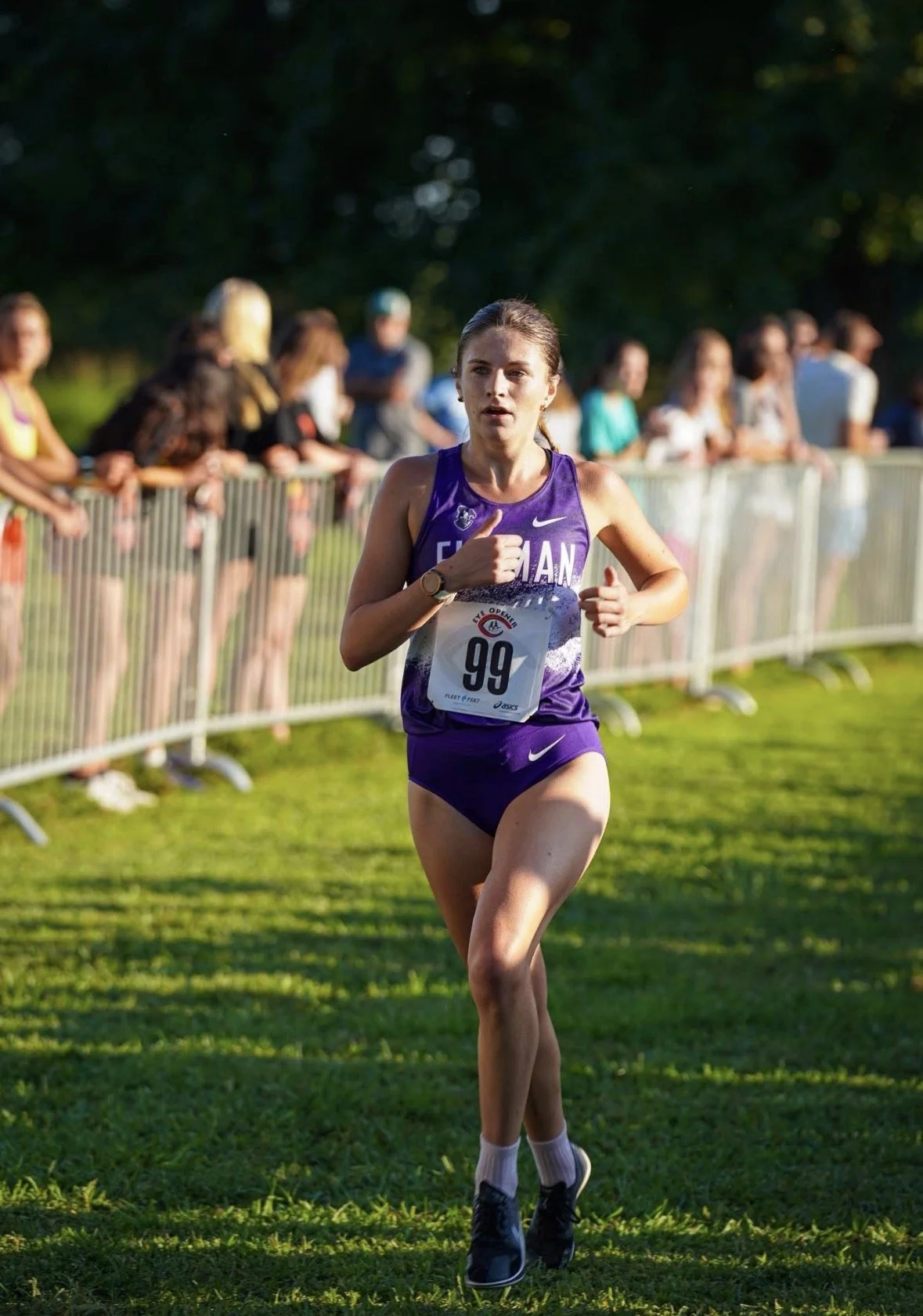A female athlete wearing a purple track uniform with the number 99 is running on a grassy field during a cross-country race, with spectators behind a fence watching her.