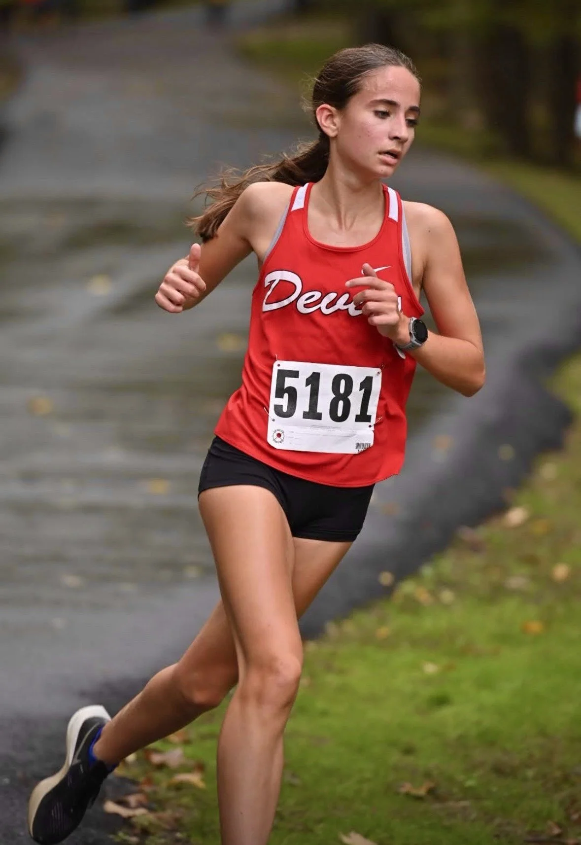 A female runner with a race bib number 5181 wearing a red tank top and black shorts, running on a paved pathway surrounded by grass and trees.