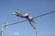 A person performing a high wire act, hanging off a tightrope in midair outdoors against a blue sky.