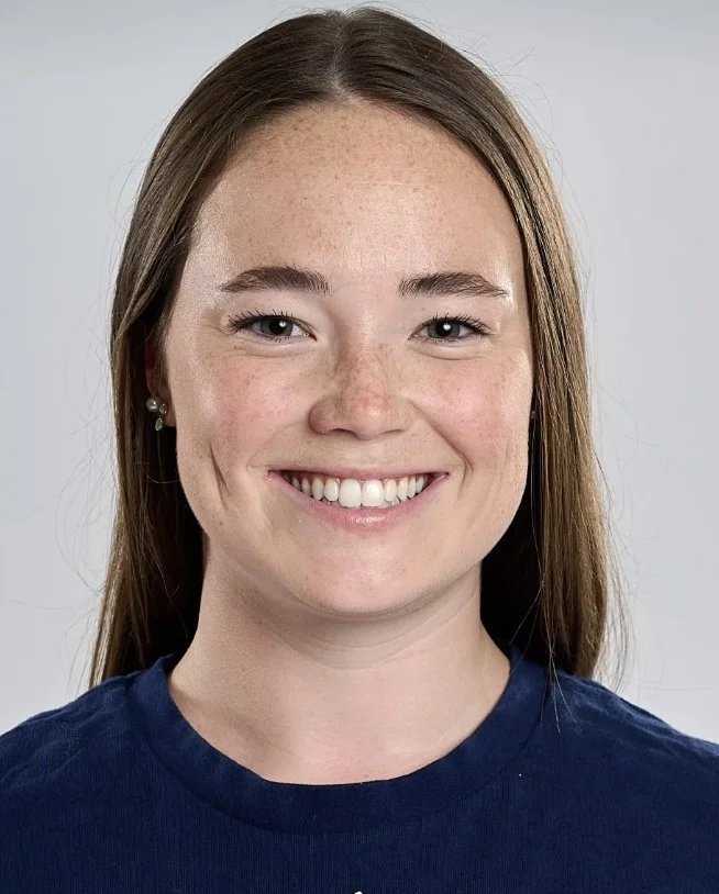 A young woman with long brown hair, wearing a dark blue shirt and earrings, smiling against a gray background.