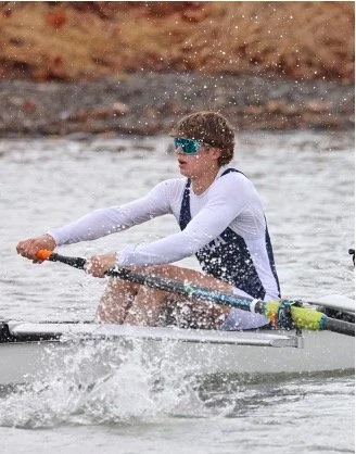 Person wearing sunglasses and a white long-sleeve shirt rowing a boat on the water.