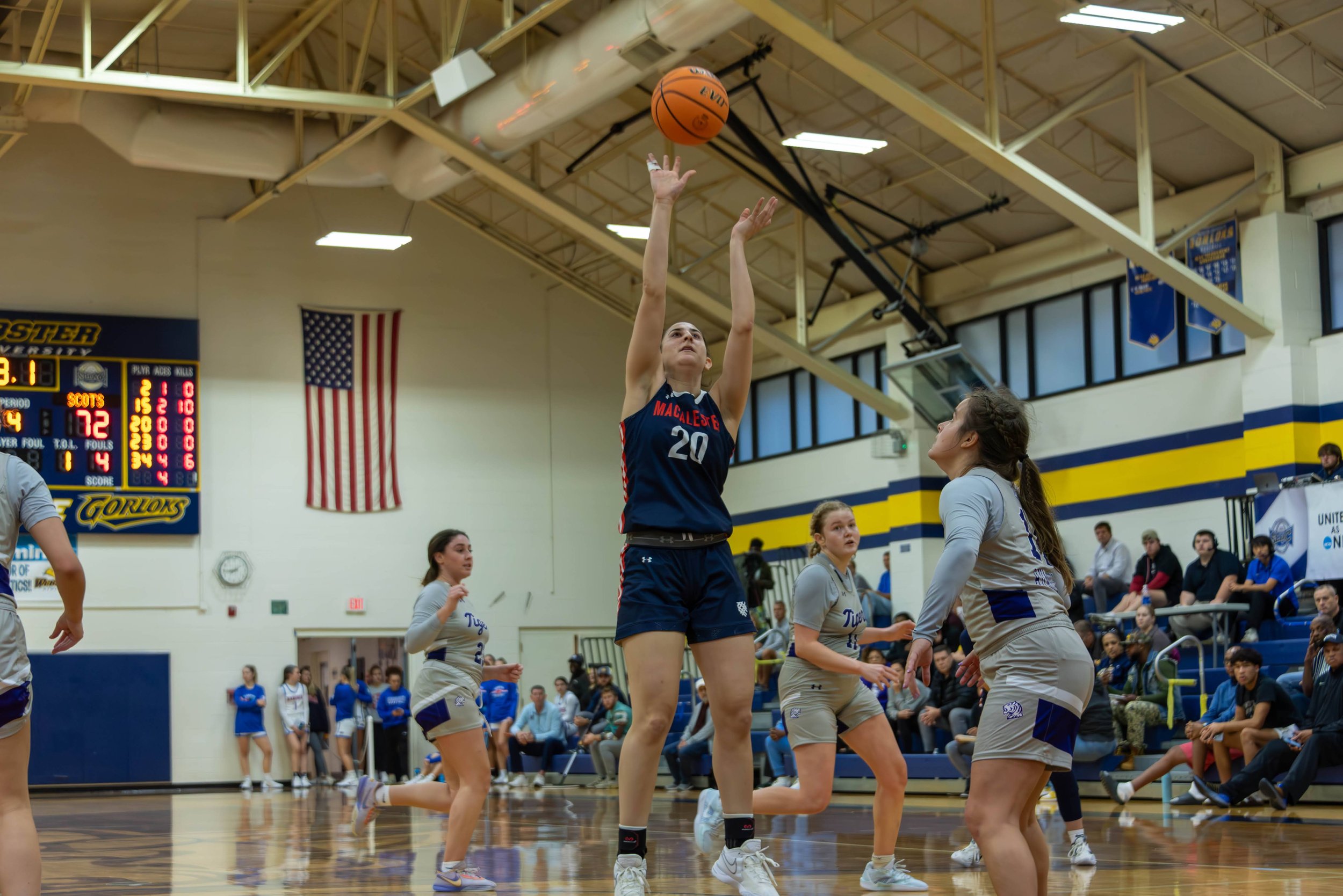 A female volleyball player wearing a navy blue and red uniform with the number 20 jumps to hit the ball during a game, while an opponent in a gray and purple uniform watches from below. The game is taking place in a gymnasium with a scoreboard, American flag, and spectators in the background.