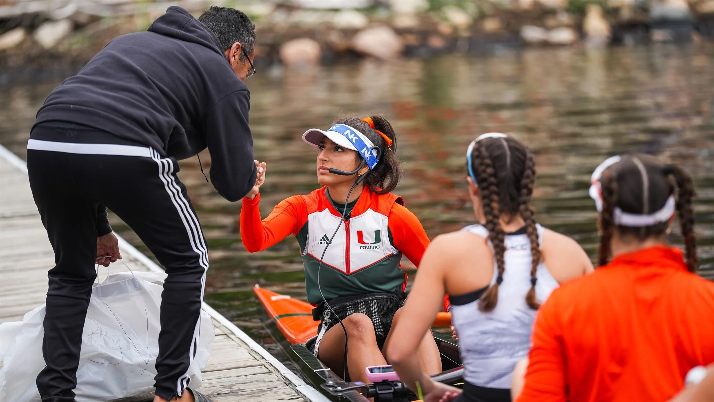 A woman in orange and black athletic gear, wearing a headset, sitting in a rowing shell near a dock, talking to a man standing outside the boat. Two other women in athletic clothing sit with her, with braided hair, near a body of water with rocks along the shore.