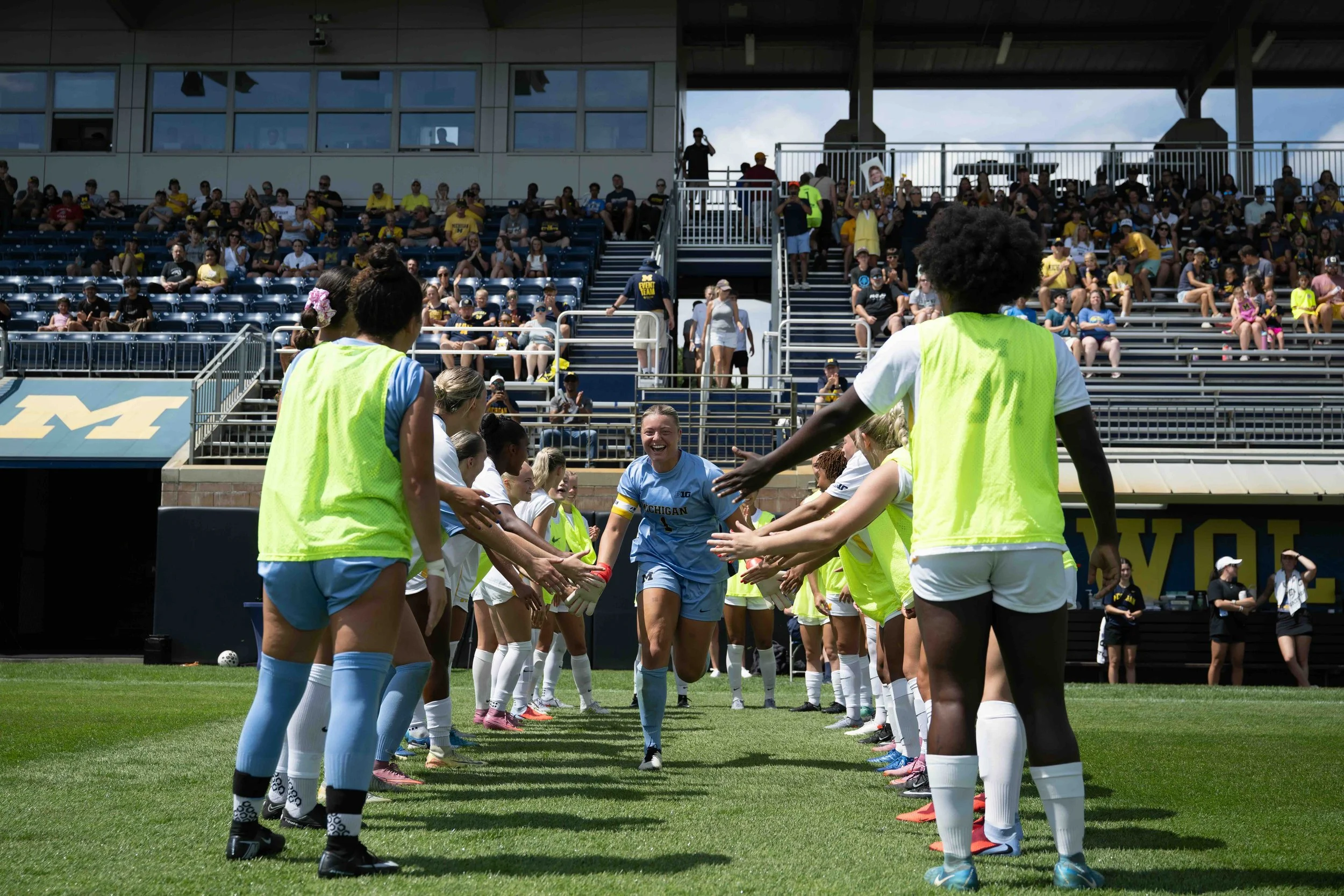 A women's soccer team in bright yellow jerseys and white shorts greeting each other on the field, while spectators watch from the bleachers in the background.