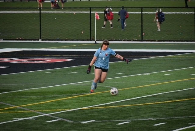 A female soccer goalkeeper in a light blue jersey and black shorts kicking a soccer ball on a field during a game.