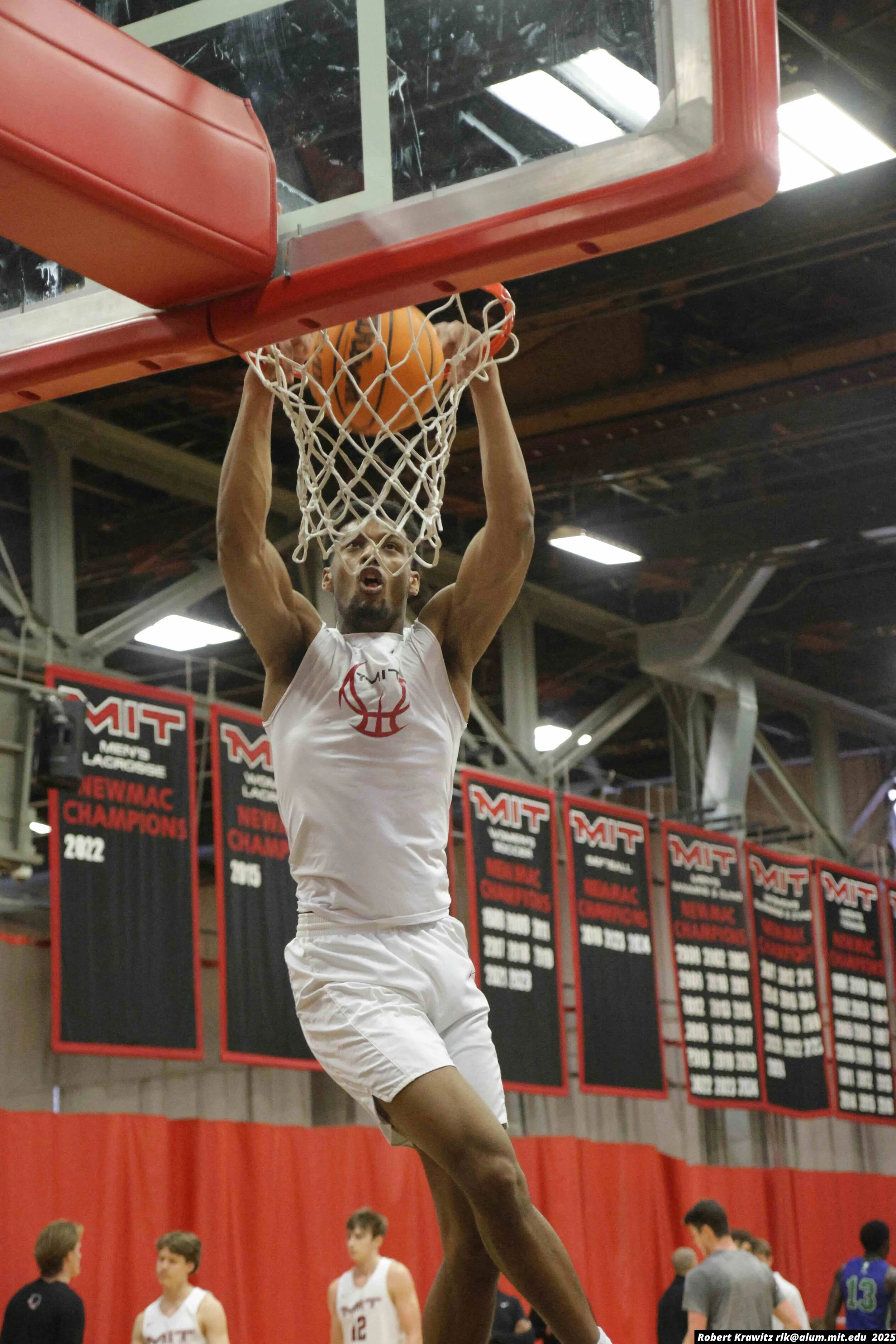 A basketball player in a white uniform is dunking a basketball into a hoop during a game at MIT. Several other players and spectators are visible in the background.