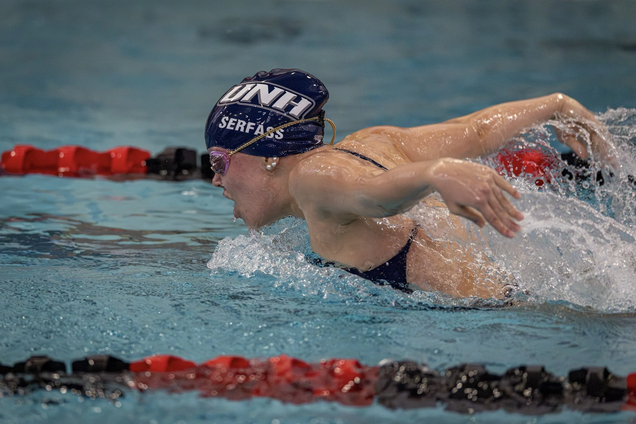 Swimmer in a swimming pool wearing a black swimsuit, a navy blue swim cap with white lettering, goggles, and a pearl earring, performing a front crawl stroke.