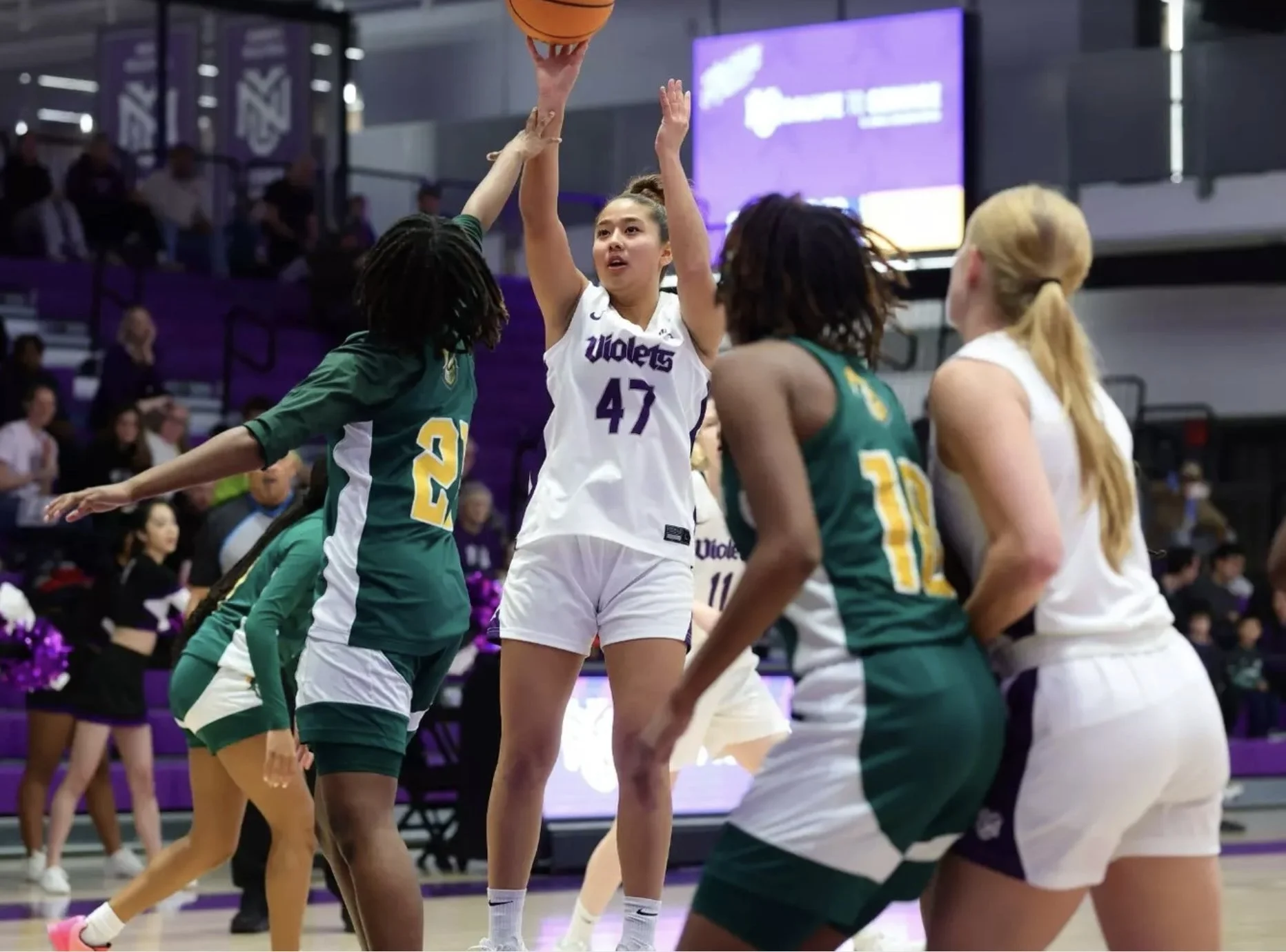 A women's basketball game in progress with a player in a white jersey numbered 47 aiming a shot at the basket, while players in green jerseys attempt to block or defend. Spectators and team staff are visible in the background.