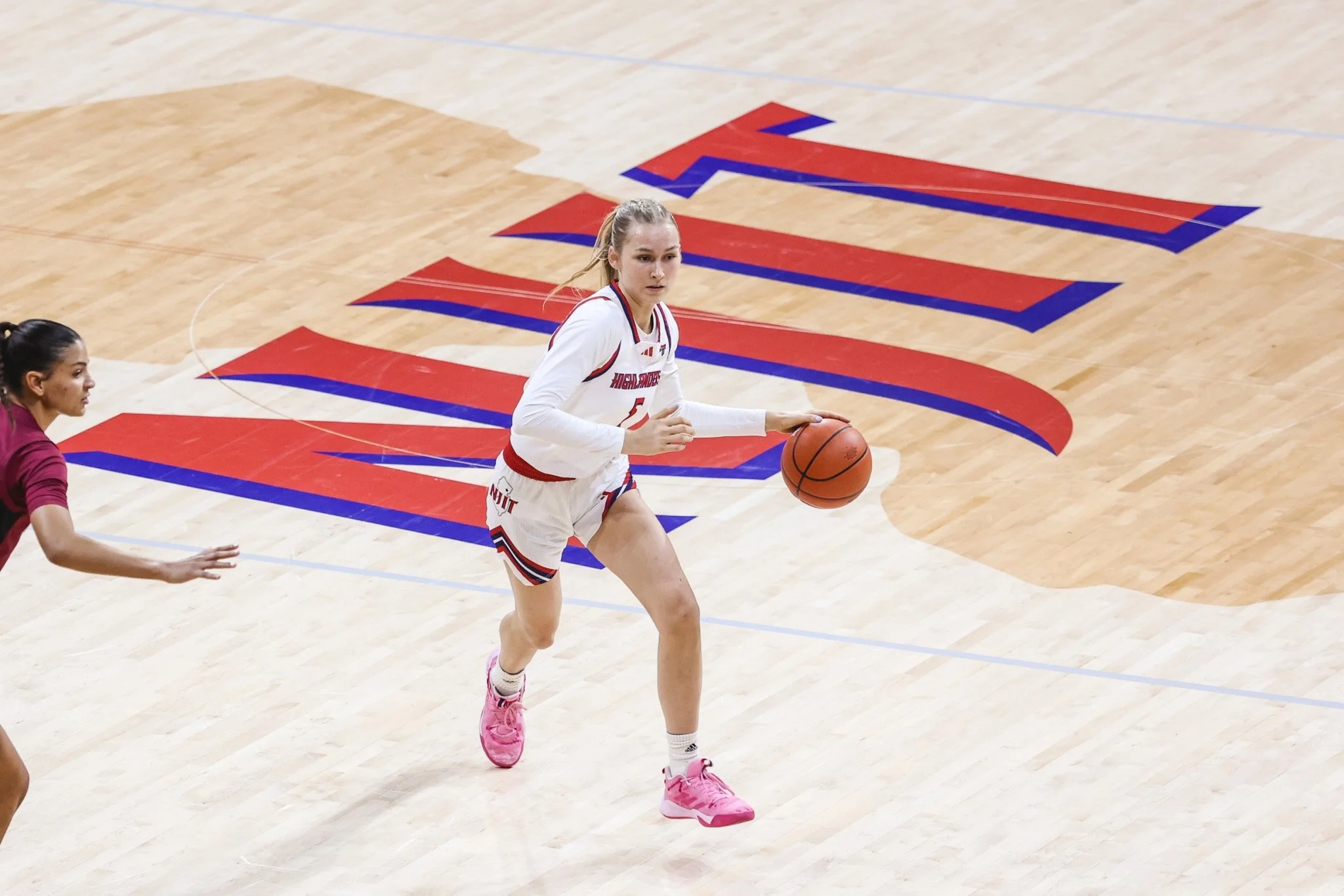 A female basketball player in a white and red uniform dribbling a basketball on a wooden court with a large logo.