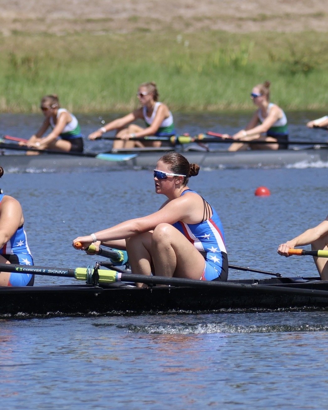Women rowers in a competitive race on a river, wearing athletic gear and sunglasses.