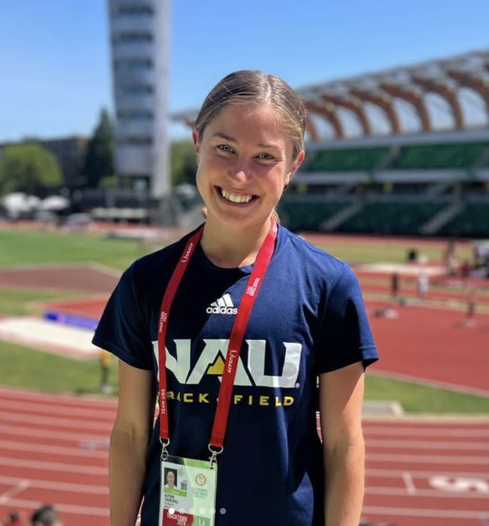 Young woman smiling at an outdoor track and field stadium, wearing a navy blue NAU track and field shirt, with a red medal around her neck and an ID badge hanging from a red lanyard.