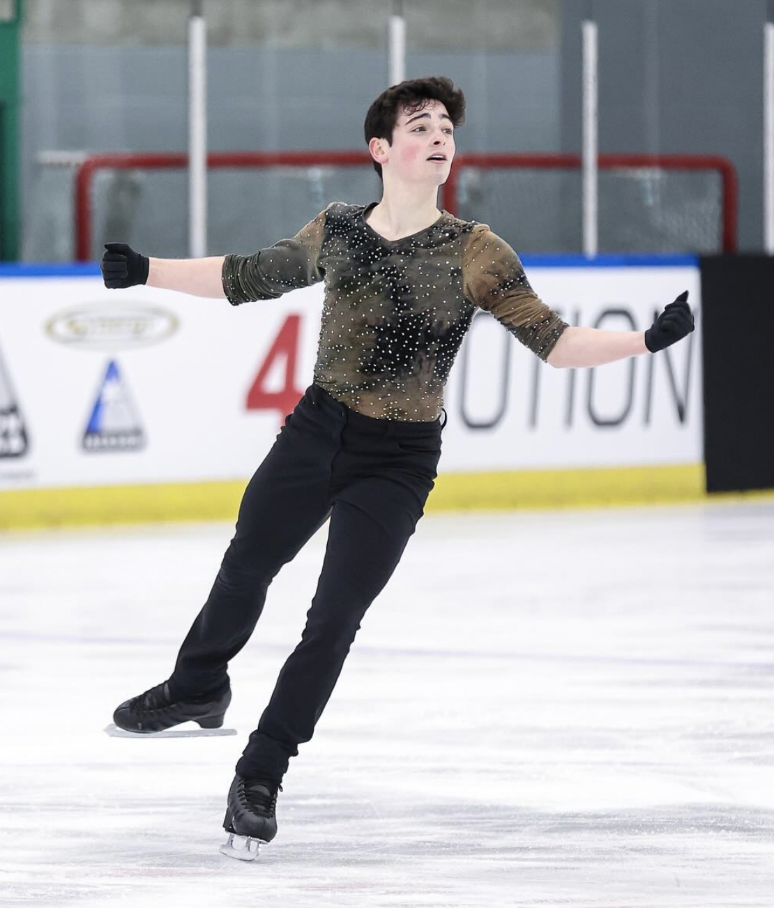 A male figure skater gliding on ice rink, wearing black pants, a sheer, embellished top, and black gloves, with arms outstretched and a focused expression.