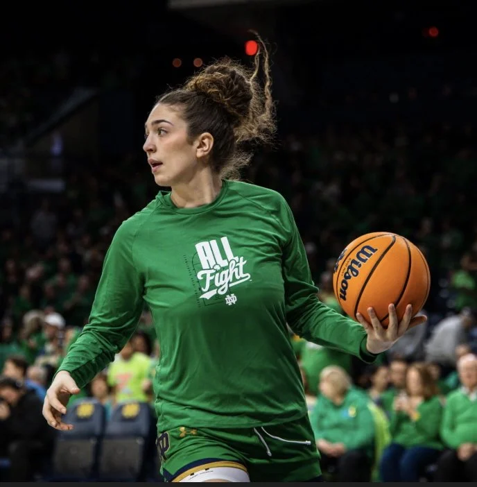 A woman with long brown hair in a bun, wearing a green sports shirt and shorts, holds a basketball during a game in an indoor arena with spectators in the background.