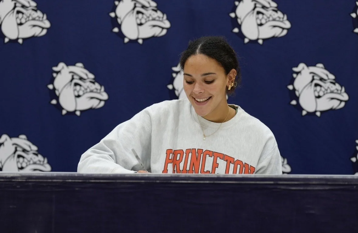 A young woman with dark hair and gold jewelry, smiling while signing a document at a table, with Princeton Bulldogs logos on a blue background behind her.