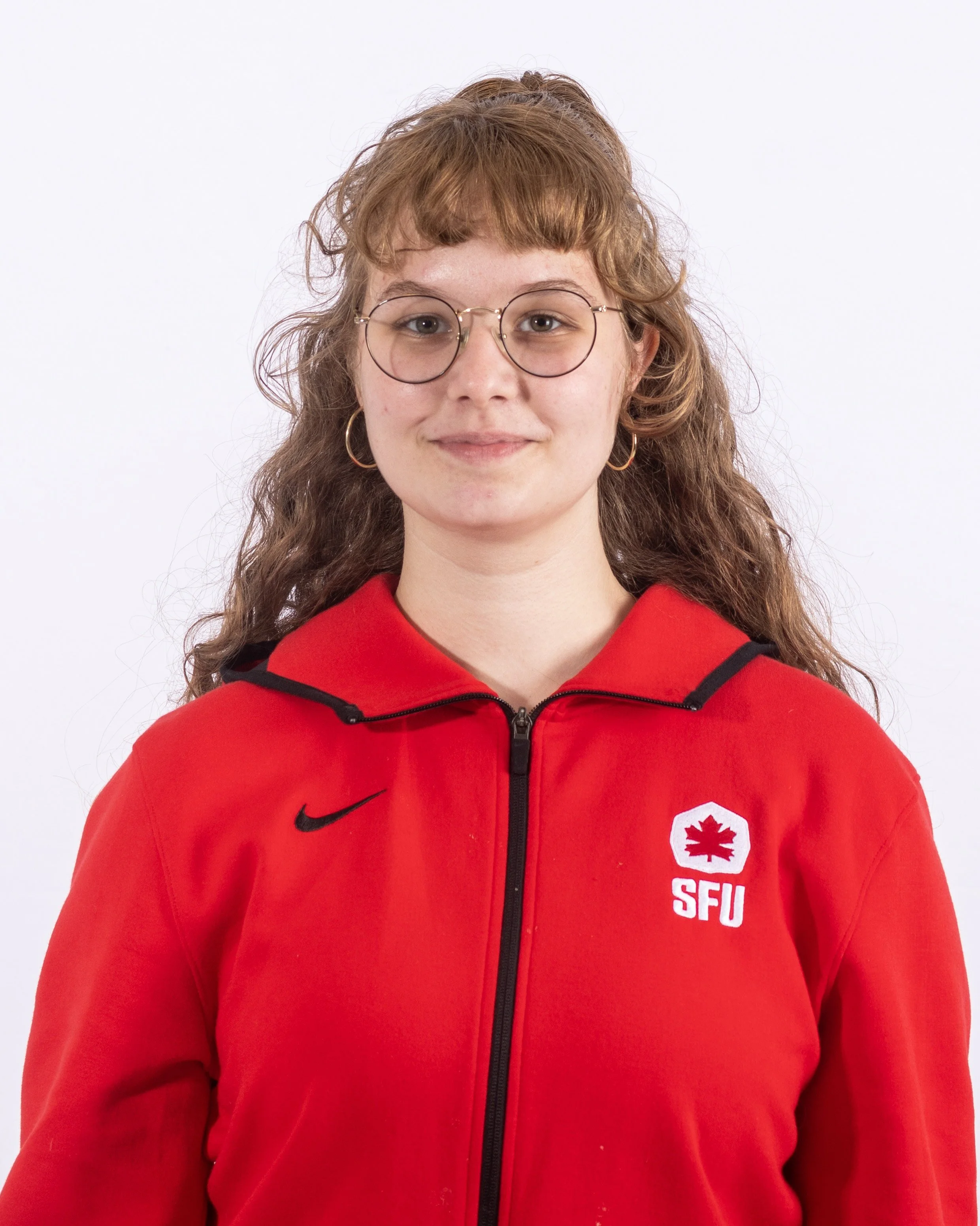 A young woman with curly reddish hair, wearing glasses, a red hoodie with a black Nike logo and a white shield emblem with a red maple leaf and the letters 'SFU' on the chest, stands against a plain white background.