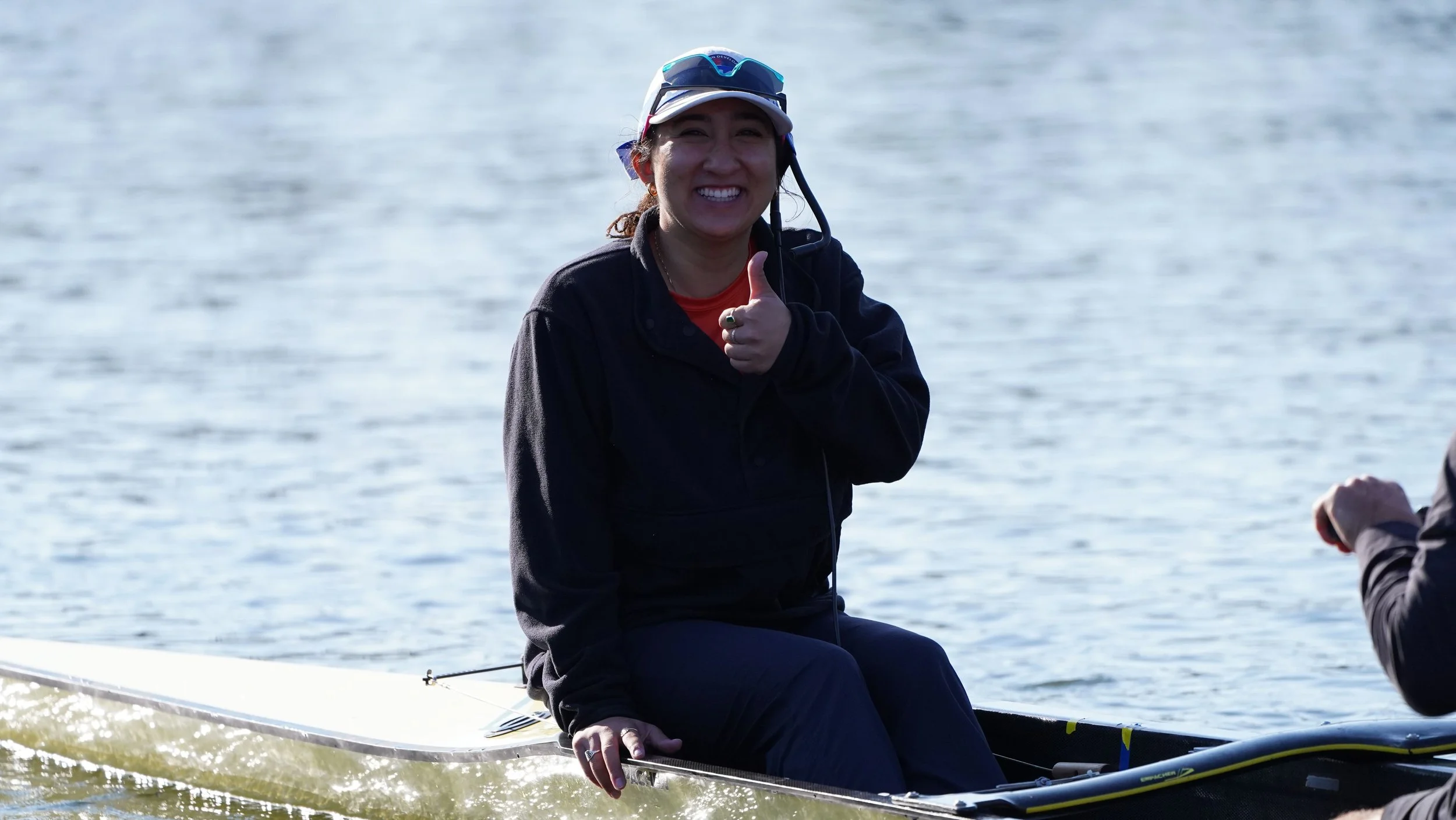 A woman smiling and giving a thumbs-up while sitting on a boat on the water, wearing a life vest and sunglasses.