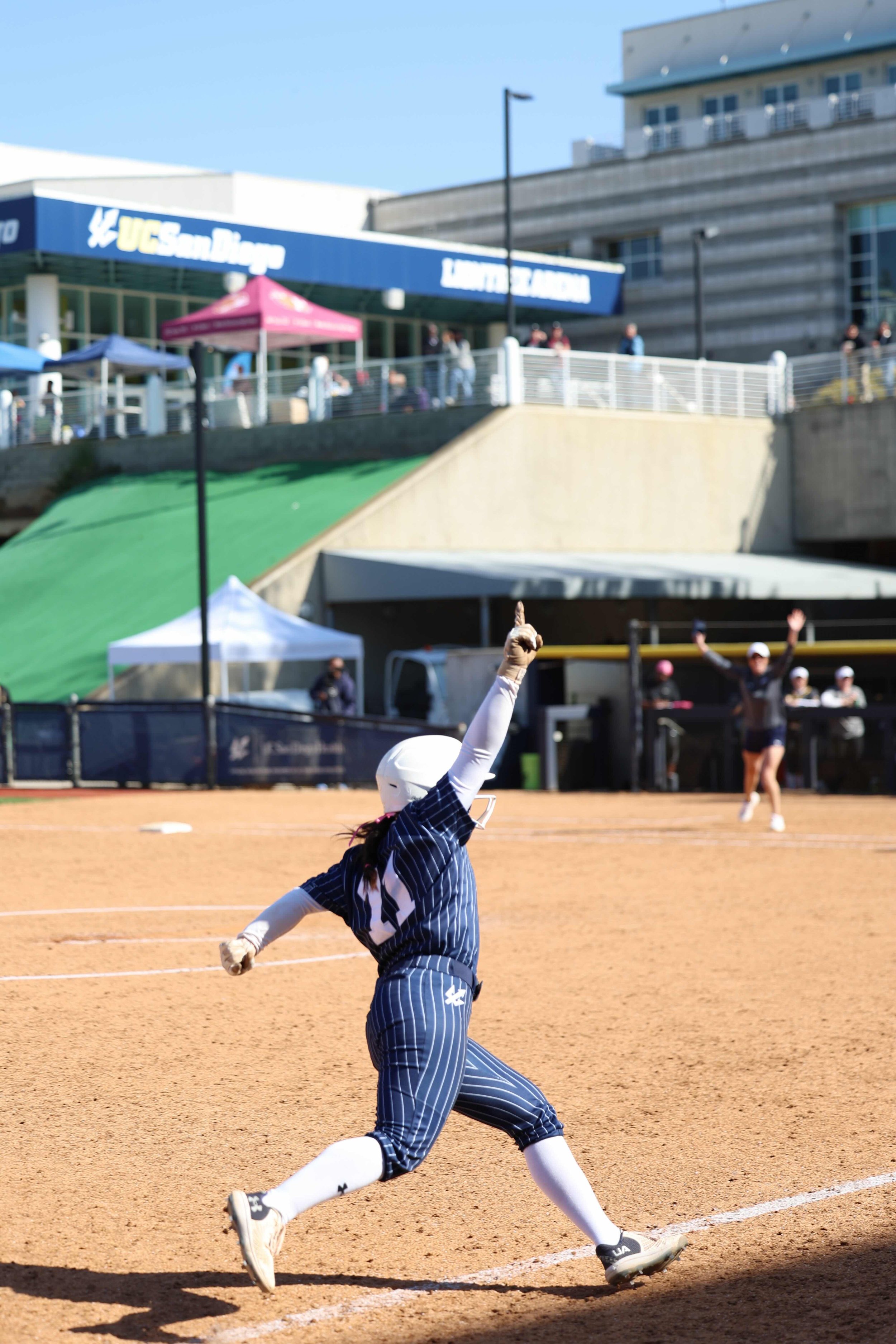 A young girl in a striped blue and white softball uniform sliding into third base during a game at UC San Diego. She is wearing a white helmet, white gloves, and white cleats, and is reaching out with her right arm as she slides on the dirt field. Other players and spectators are visible in the background near the stadium.