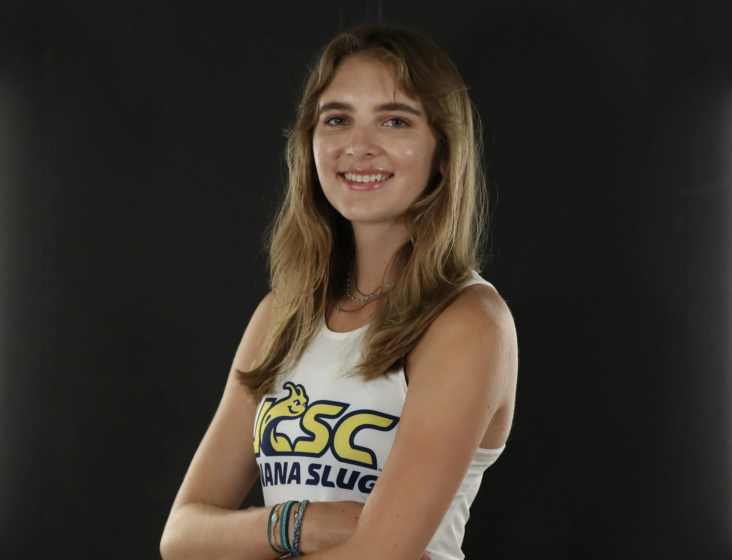 Young woman with long hair smiling, wearing a sleeveless white shirt with text and a yellow graphic, standing against a black background.