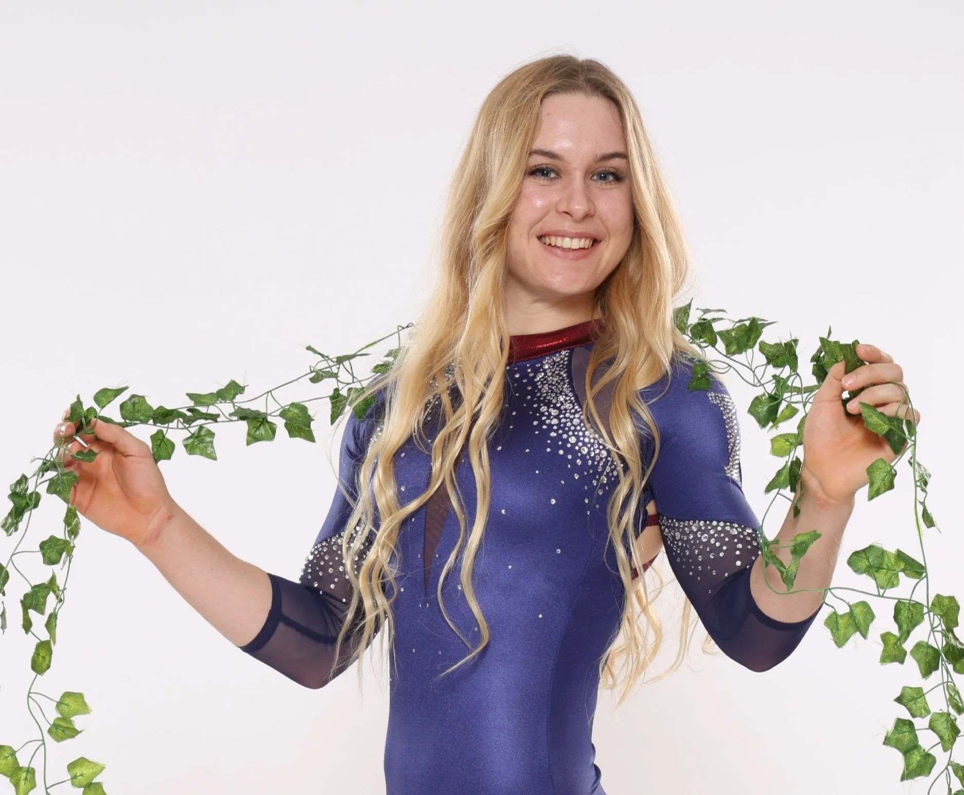 Young woman with long blonde wavy hair wearing a dark blue dress with rhinestones, smiling, holding green leafy vines against a white background.