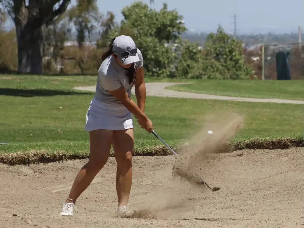 A woman playing golf in a sand bunker, preparing to hit the golf ball with a wedge.