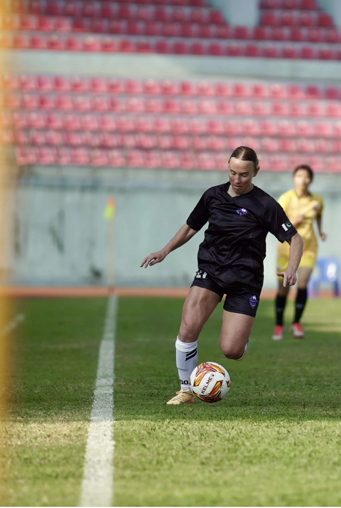 A female soccer player in black uniform preparing to kick a soccer ball on the field during a match in a stadium with pink empty seats.