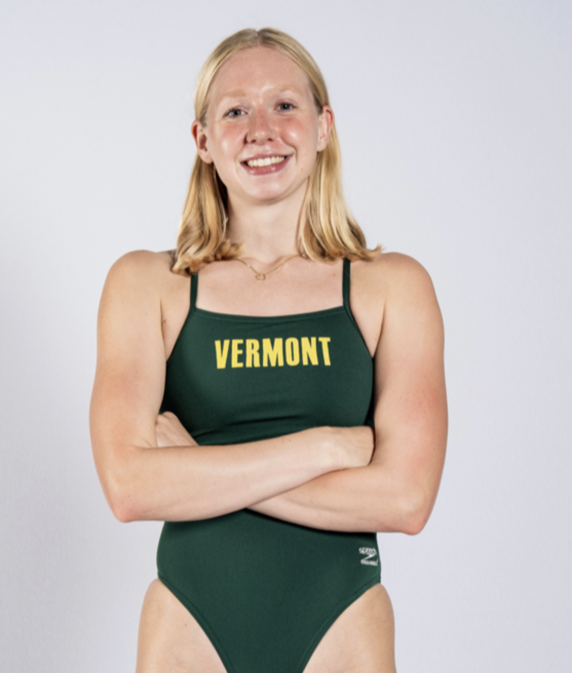 A young woman with blonde hair, smiling, wearing a green athletic swimsuit with yellow lettering that says 'VERMONT' and crossed arms.